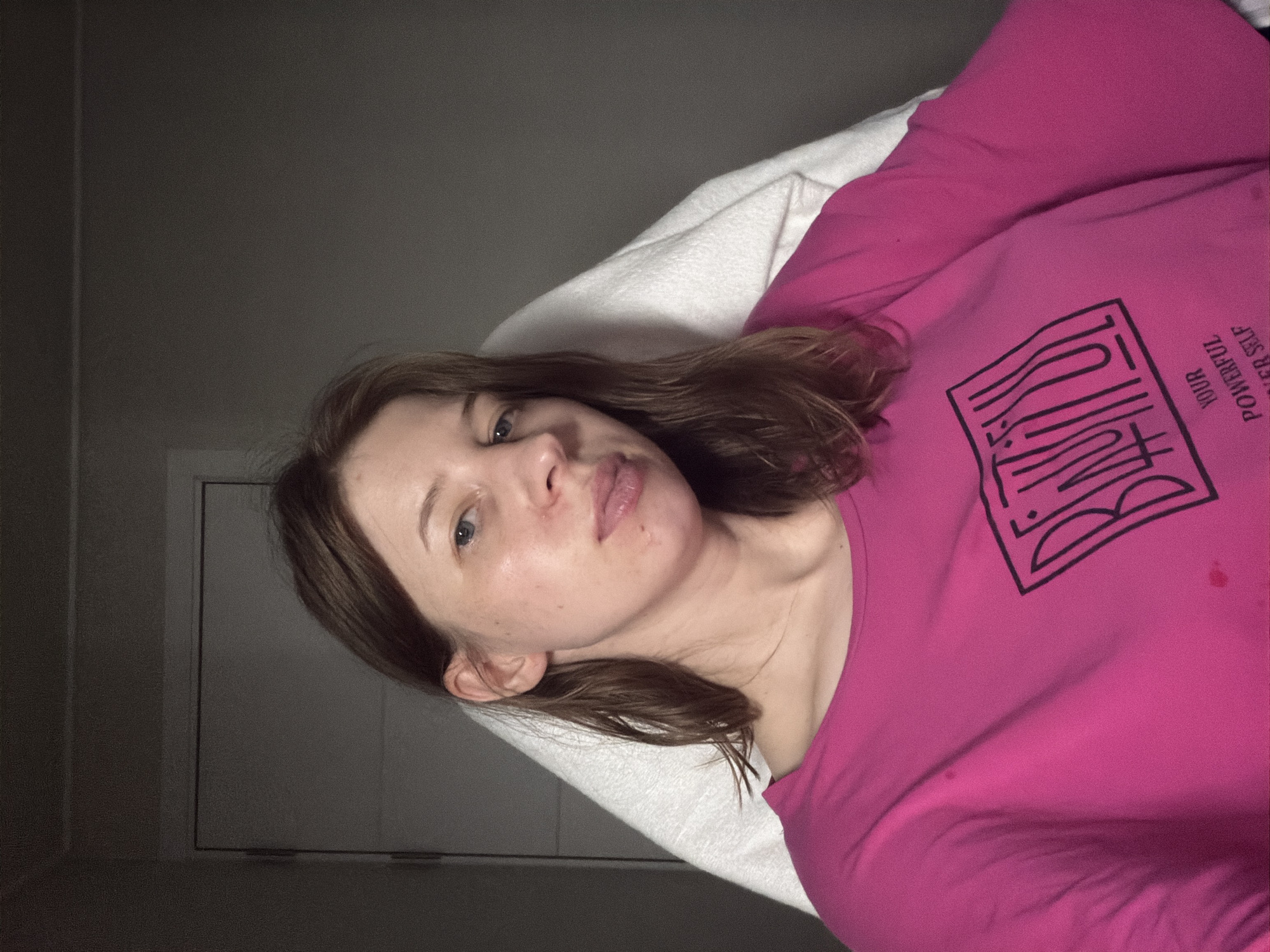A woman with brown hair is sitting in a chair, leaning against a white blanket. She is wearing a bright pink t-shirt with a black graphic design that appears to say "BEAUTIFUL" in stylized lettering, followed by smaller text that reads "YOUR POWERFUL INNER SELF." The background consists of a grey wall and a white door. The lighting suggests an indoor setting, possibly a home or a room, with the overall ambiance being relaxed and casual. There are no other individuals present in the frame. The scene does not contain any elements that specifically place it in Dongducheon-si, South Korea.