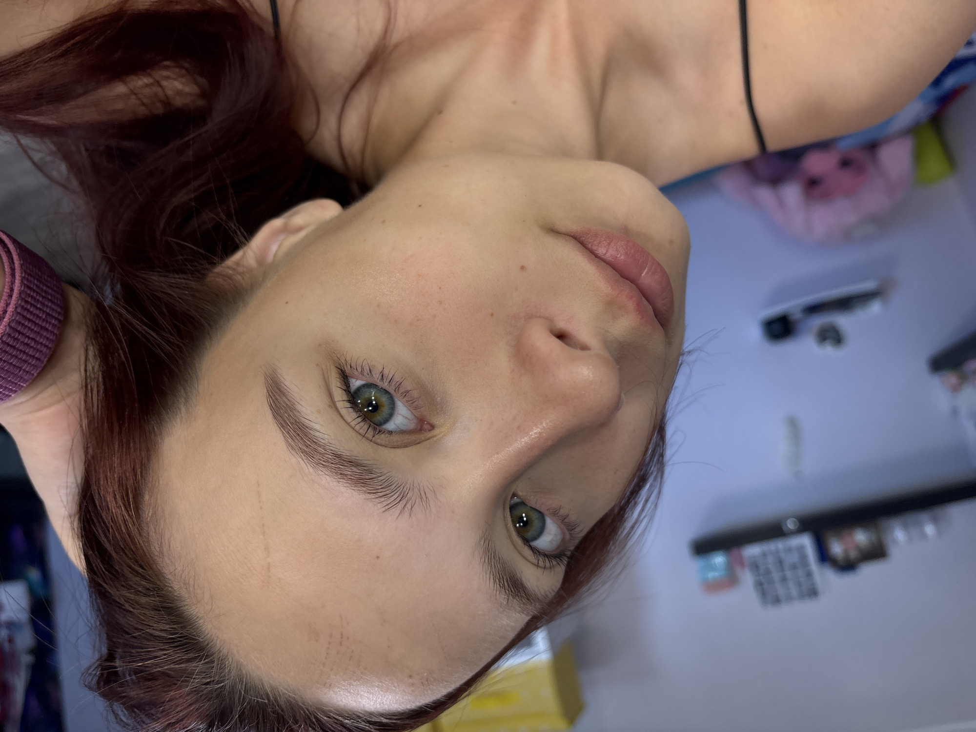 This is a close-up, high-angle portrait of a person, likely female, with fair skin, striking green/hazel eyes, and reddish-brown hair. The subject is lying down, looking directly upwards towards the camera with a neutral, calm expression. Their hair is spread around their head, framing the face, with some strands visible on the forehead.
Visible features include well-defined eyebrows, long eyelashes, and slightly parted lips. A few small moles or freckles are present on the skin. The person is wearing a thin black strap, likely from a top, visible near the neck/shoulder area, and a purple ribbed band on their left wrist or arm.
The background is largely out of focus, suggesting an indoor setting. Hints of light-colored bedding or a wall are visible, along with a blurred, patterned fabric (pink and blue) in the upper right. Further back, indistinct shapes, including a horizontal dark object (possibly a shelf or furniture) and a blurred, grid-like pattern resembling a QR code, are discernible but illegible. The image conveys a direct and intimate gaze from the subject. The setting is an indoor space in Прикамский, Russia.