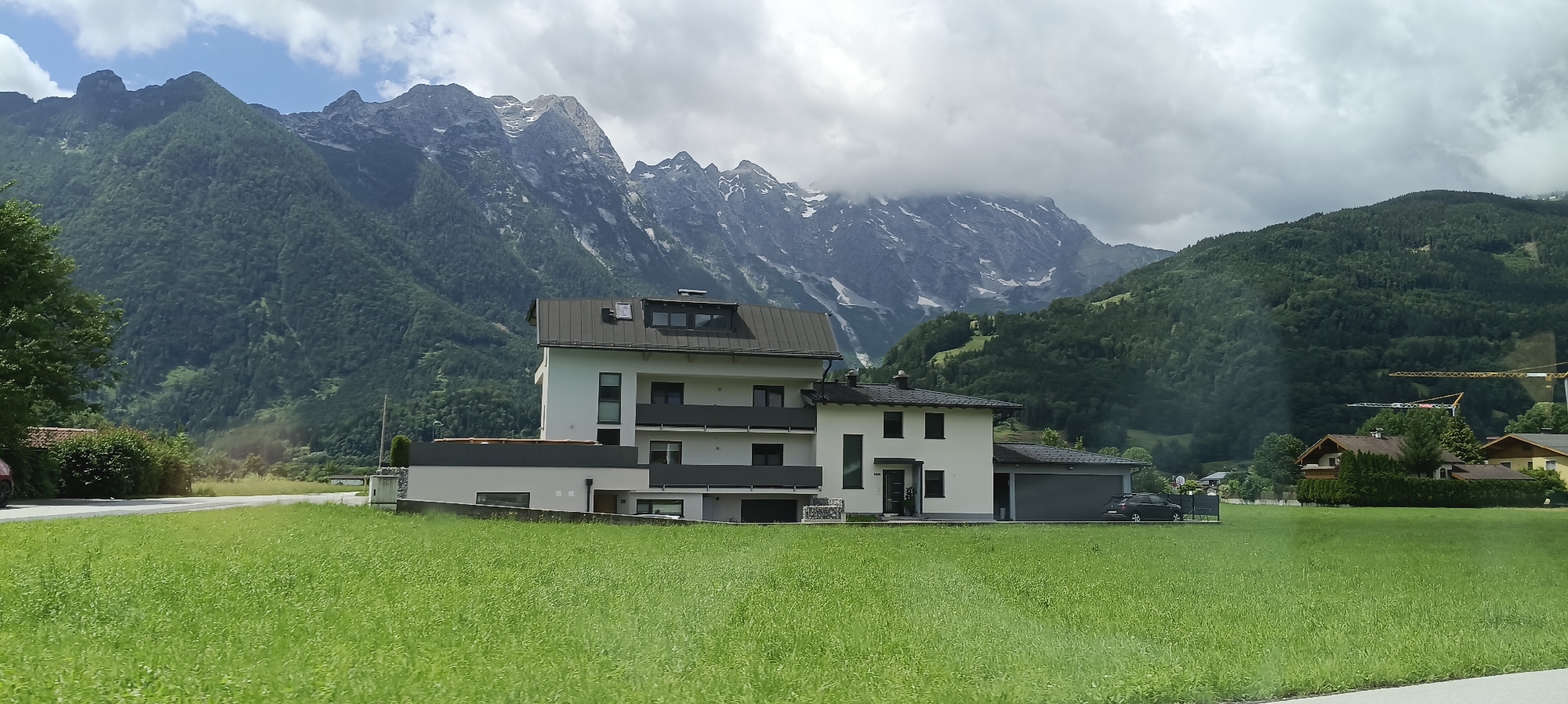 In Kellau, Austria, a modern white house with dark gray balconies and a dark gray roof stands prominently in the foreground. The house features multiple stories, visible windows, and a garage. To the left of the house, a green lawn extends to a road, with a dark-colored vehicle parked near the garage. Further to the left, a dense green bush and a portion of a red-tiled roof are visible. Behind the house, a vast, lush green mountain range dominates the background, with a cloudy sky overhead. Snow-capped peaks are visible on the higher elevations of the mountains. The overall environment suggests a rural or semi-rural setting within a mountainous region.