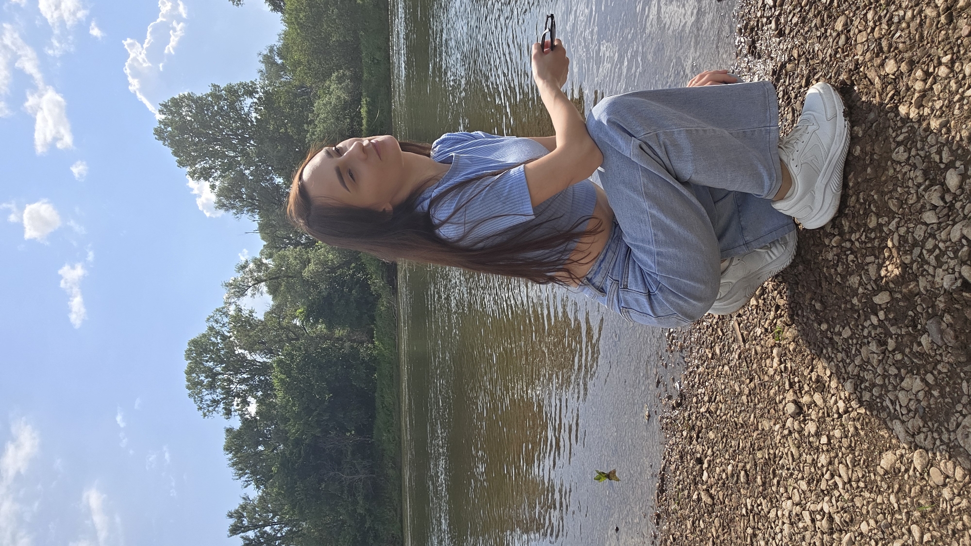 A young woman with long, dark hair is crouching on a rocky shore beside a river. She is wearing a light blue ribbed crop top and light blue wide-leg jeans, paired with white sneakers. Her head is tilted back, and her eyes are closed, as if basking in the sunlight. In her right hand, she holds a pair of dark-rimmed glasses. The river behind her is calm, reflecting the trees and the blue sky with scattered white clouds. The shoreline is covered in pebbles and small rocks, with a few blades of grass peeking through. The background consists of a dense line of green trees. The scene appears to be a natural outdoor setting.