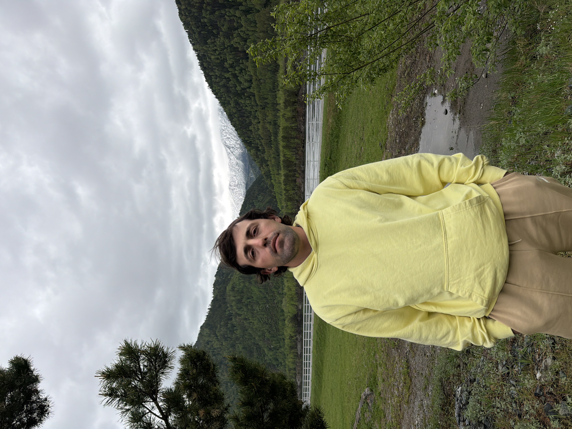 A man wearing a yellow hoodie and tan pants stands in a field with mountains in the background. The sky is cloudy. There are green trees and grass visible in the field. There is also a small pond or puddle visible. The mountains are covered with green trees and some snow. The man appears to be in a natural outdoor setting. There are no clear markers to identify the exact location in Cheremushki, Russia.