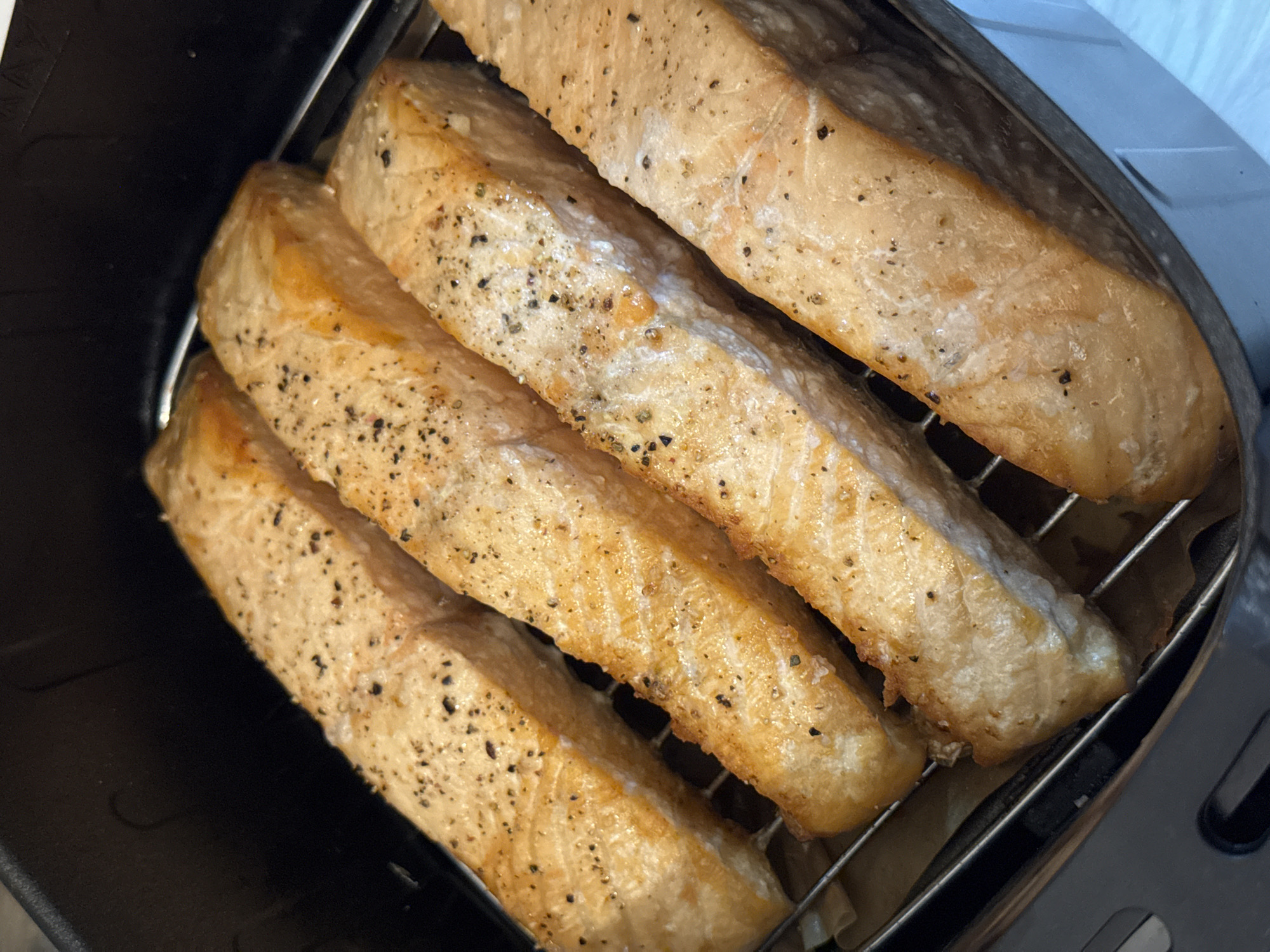 The image shows four cooked salmon fillets arranged side-by-side on a wire rack inside what appears to be an air fryer. The fillets are seasoned with black pepper. The scene appears to be indoors. The location in the city of Chaikovsky, Russia is not visible in the image.