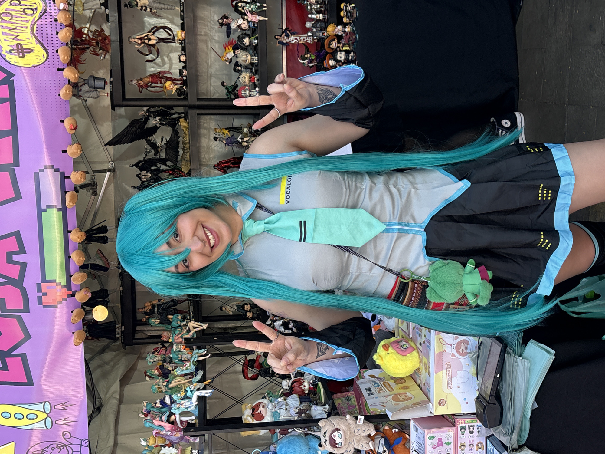 A young woman with bright teal hair is posing in front of a display of anime figurines at an outdoor market. She is wearing a blue and gray Vocaloid costume, complete with a teal tie and a black skirt. She is holding up two fingers in a peace sign. The background shows shelves filled with various anime figurines, and a purple banner with yellow and pink lettering is visible above. The scene appears to be at an anime or comic convention, potentially in a city like São Paulo, Brazil, given the prevalence of such events. The atmosphere is vibrant and colorful, with many toys and collectibles on display.
