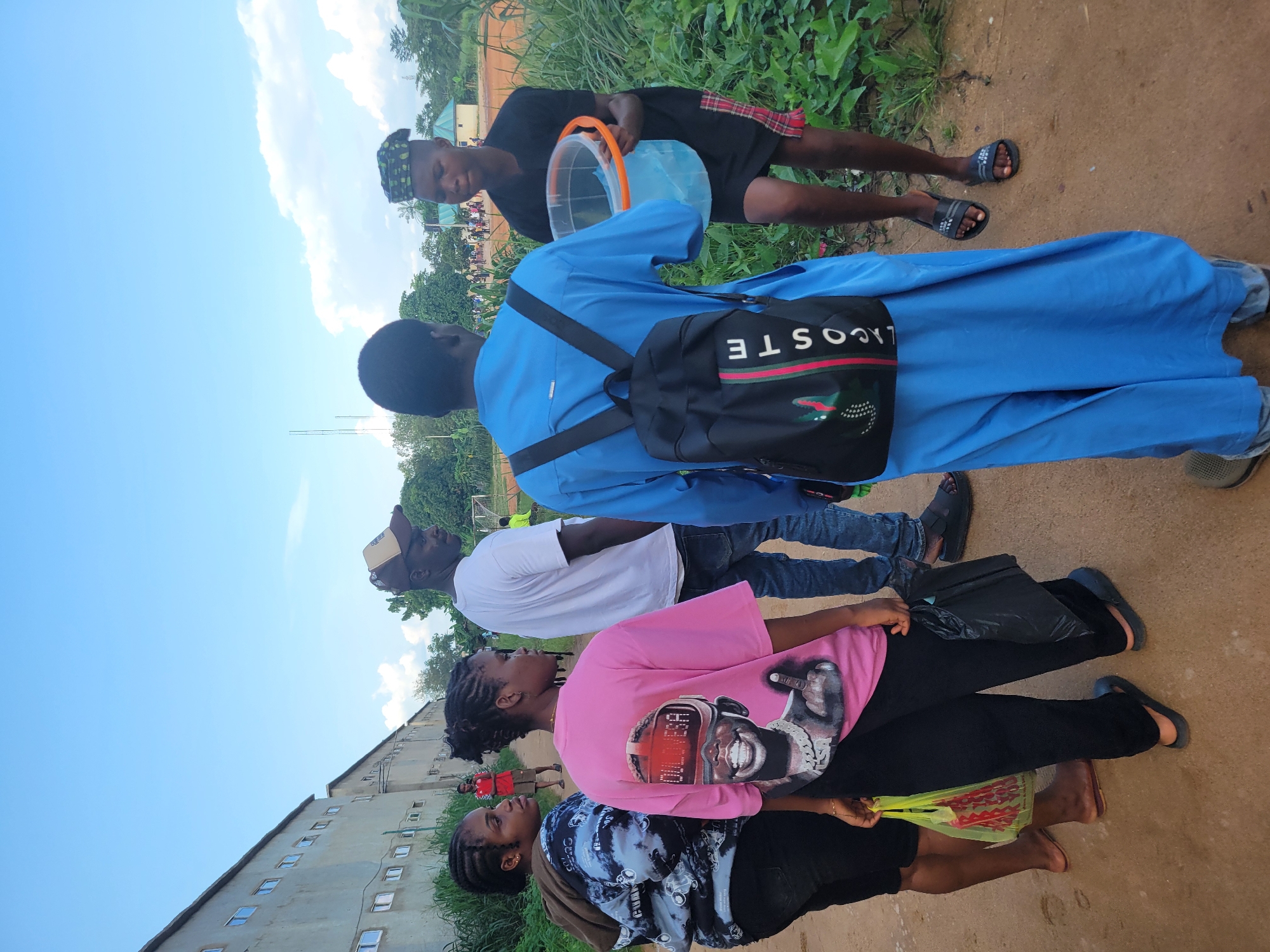 A group of people stand on a dirt path, with a building and lush green vegetation in the background. The sky is a clear blue with some white clouds, suggesting it is daytime with fair weather. One person in the foreground wears a blue robe and a black backpack with the "LACOSTE" logo visible. Another person behind them wears a white t-shirt. Further down the path, a person in a pink t-shirt with a graphic print of a person's face and the text "FOR REAL" is visible. Another person in a black and blue tie-dye hoodie is also present. The person in the blue robe is holding a clear plastic bucket with a blue liquid. The scene appears casual, with people dressed in everyday clothing. The presence of the building, which looks like a multi-story structure with many windows, and the natural vegetation suggest an urban or semi-urban environment. The overall mood appears relaxed, with no indication of any urgent activity.
