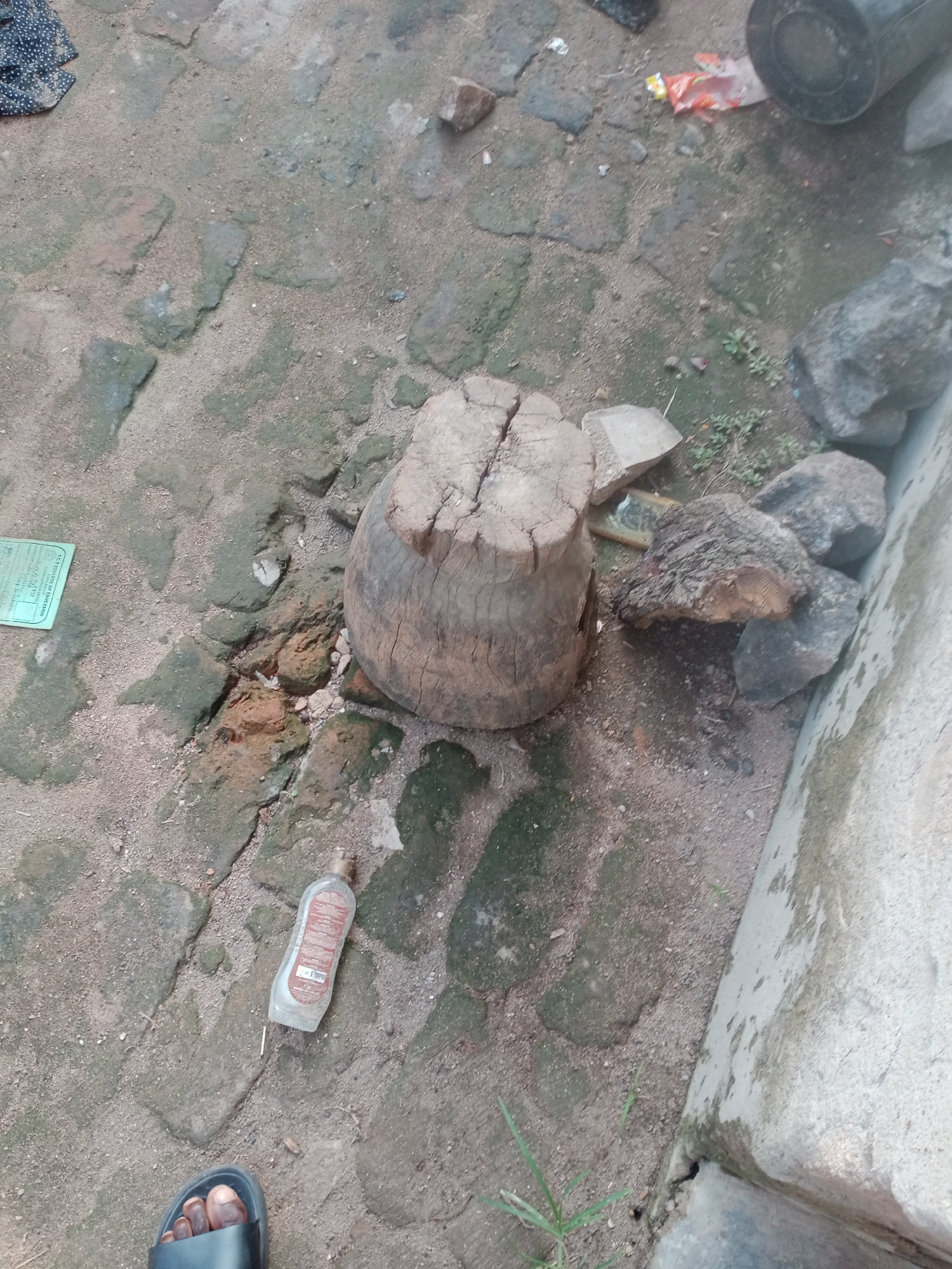 The image depicts an outdoor scene on a dirt and brick ground, likely in Jalingo, Nigeria. A rough-hewn wooden log, split at the top, stands prominently in the center. To the right of the log are several large grey rocks and a piece of wood. In the lower right quadrant, a person's foot wearing a black sandal is visible, with dark skin and painted toenails. A clear plastic bottle, possibly containing liquid, lies on the ground near the foot. Further to the left, a small green paper document is situated on the ground. The environment appears unkempt, with scattered debris and patches of greenery. The overall impression is one of an informal, possibly utilitarian, outdoor space.