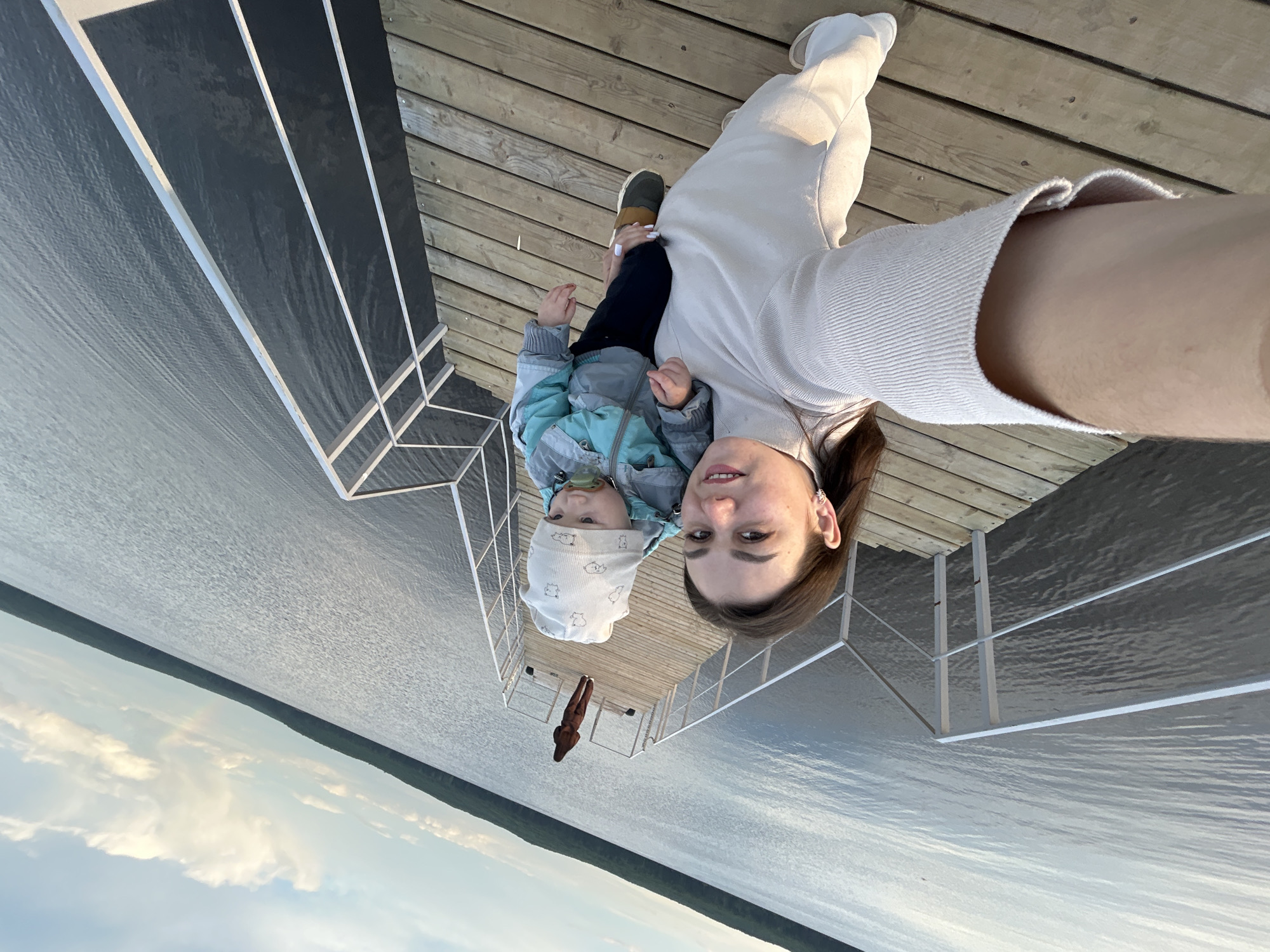 A woman holds a young child on a wooden dock, likely on the Votkinsk Reservoir in Chaikovsky, Russia. The woman is fair-skinned and wears a light-colored sweater, with dark hair pulled back. The child is also fair-skinned, wearing a light-colored patterned hat and a blue and gray jacket. The dock has a metal railing. The reservoir is visible with trees in the background under a sky with some clouds.