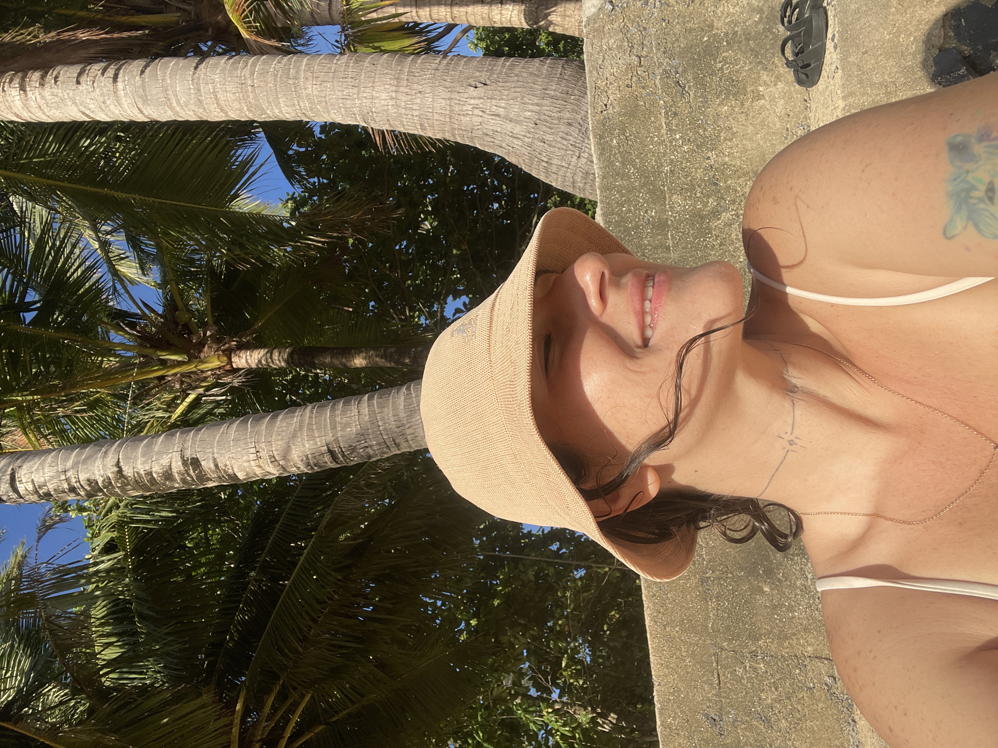 A woman wearing a light-colored sun hat and a white tank top is sitting outdoors in Rawai, Thailand, with palm trees and blue sky visible in the background. She is smiling and looking to her right. There is a concrete wall behind her, and a pair of black sandals are on the wall to her right. She has a necklace and a tattoo on her arm. The image is taken during the day with sunlight illuminating her face and body.
