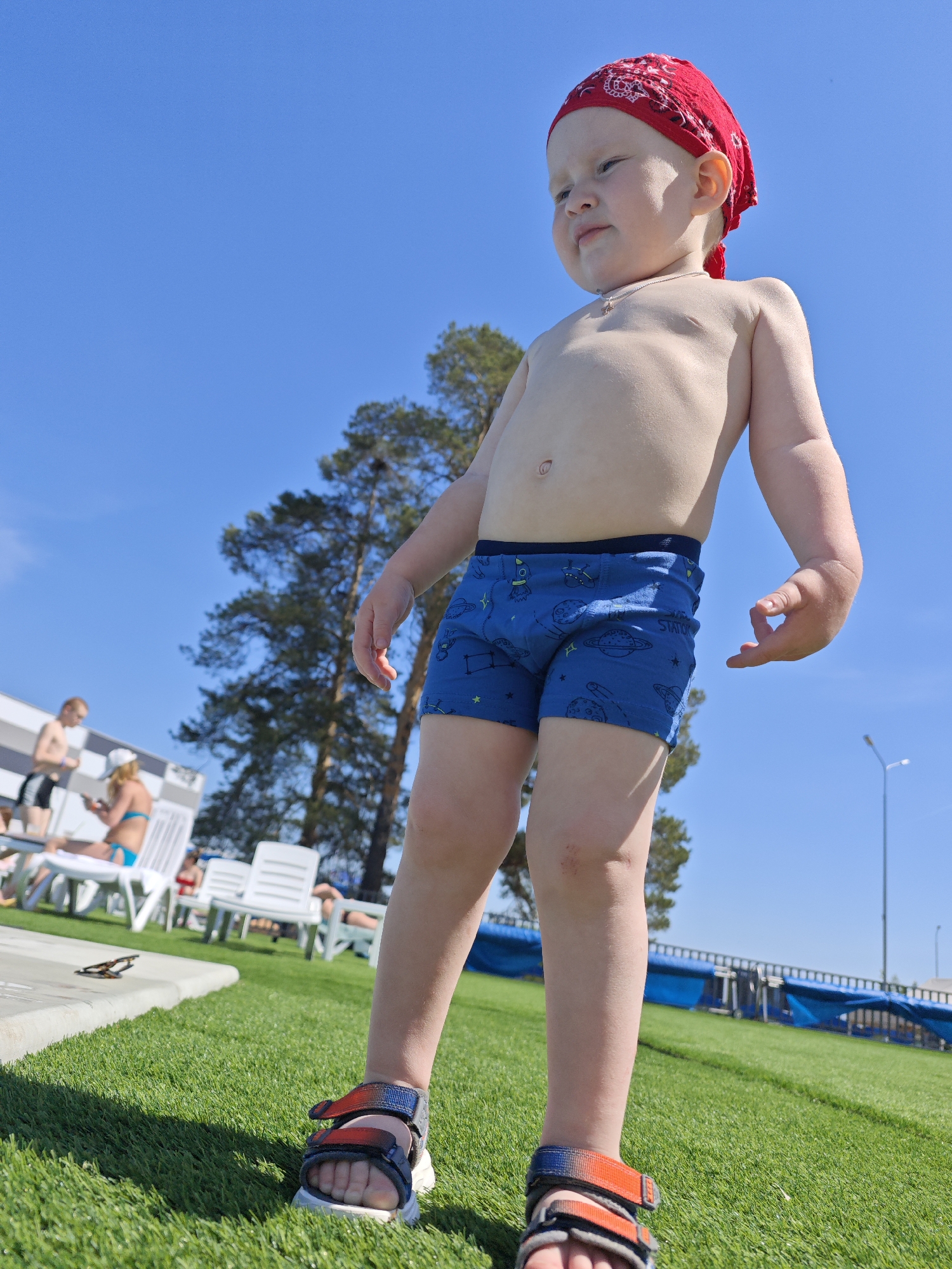 A young child wearing a red bandana, blue swim trunks with a space pattern, and sandals stands on artificial grass. The child is fair-skinned with a small necklace and is looking away from the camera. In the background, people are sitting on lounge chairs near a pool, and trees are visible against a clear blue sky. A light pole is also present in the background. The scene appears to be a recreational area, likely a swimming pool facility in Votkinsk, Russia.