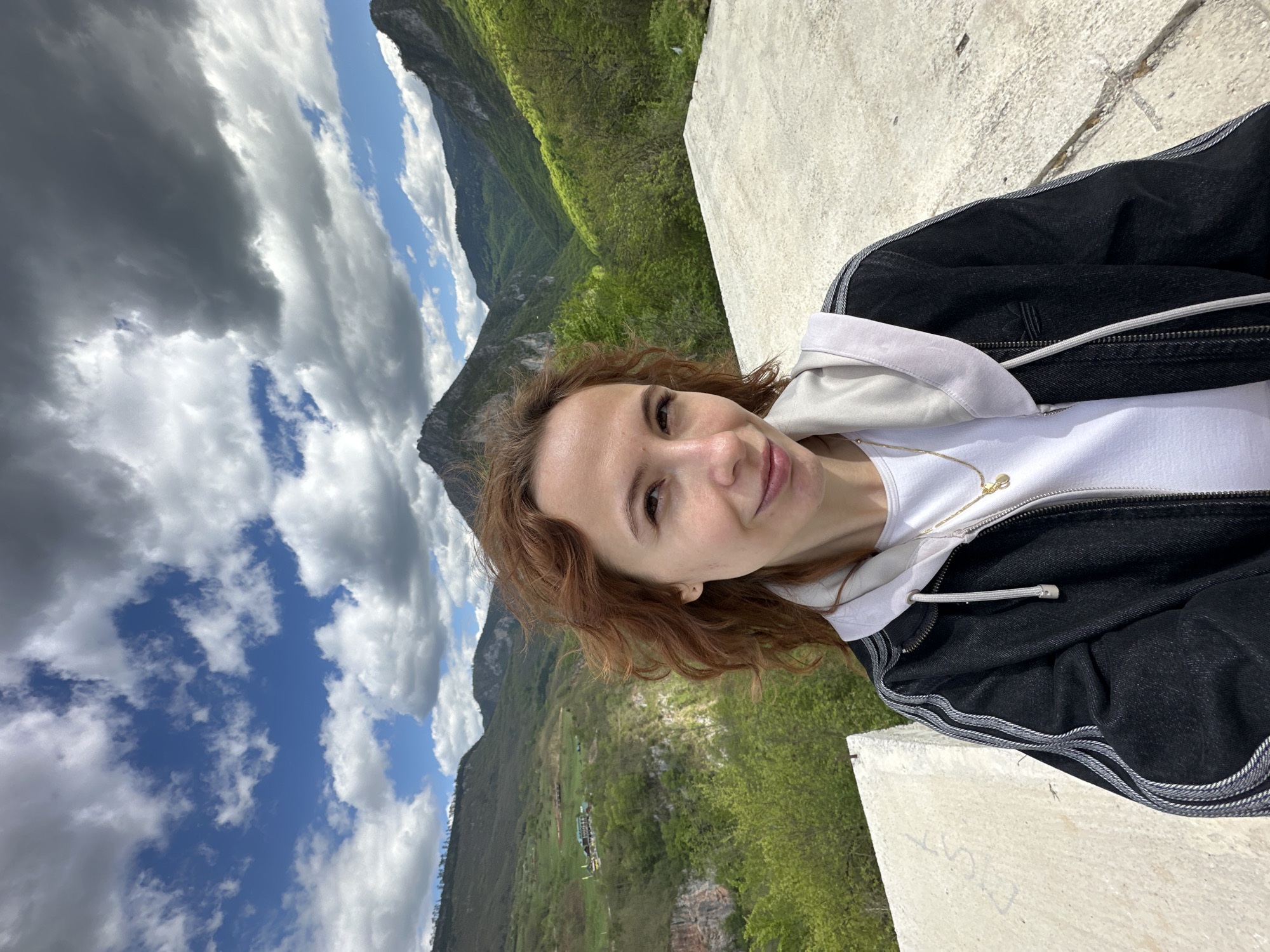 A photograph depicts a woman posing for a selfie with a mountainous landscape in the background. The woman has fair skin and reddish-brown curly hair and is wearing a white t-shirt, a light gray hoodie, and a black jacket. She is standing on a concrete platform or wall. The background features a valley with green hills and a mountain range under a partly cloudy sky. Based on the landscape, specifically the deep valley and mountain range, this image was likely taken in the area of Đurđevića Tara, Montenegro.