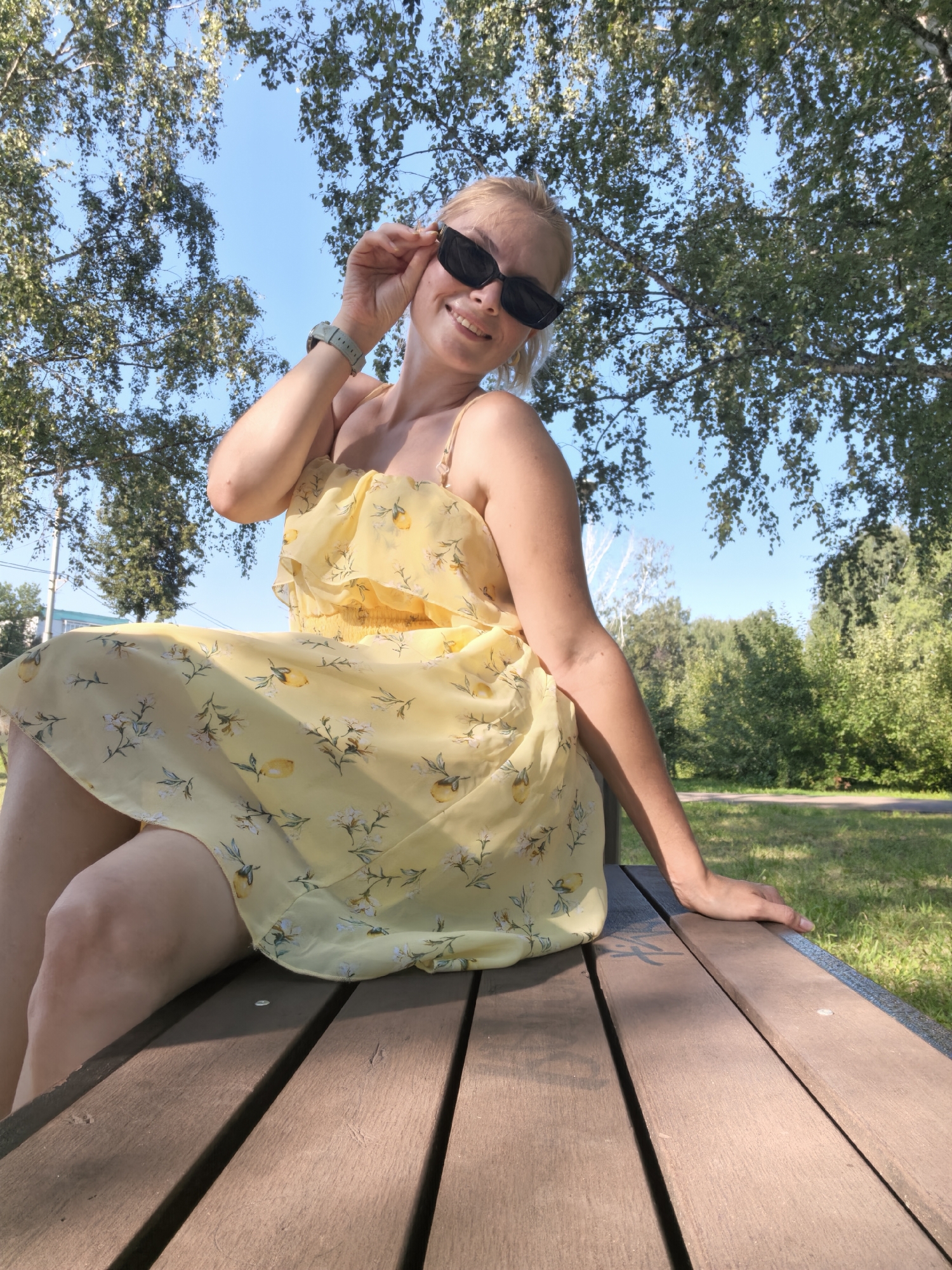 A young woman is sitting on a wooden park bench outdoors, in Sterlitamak, Russia. She is wearing a yellow dress with a floral print. She is wearing sunglasses and smiling. The background consists of trees and a clear blue sky. The bench is made of wooden planks. The image is taken from a low angle.
