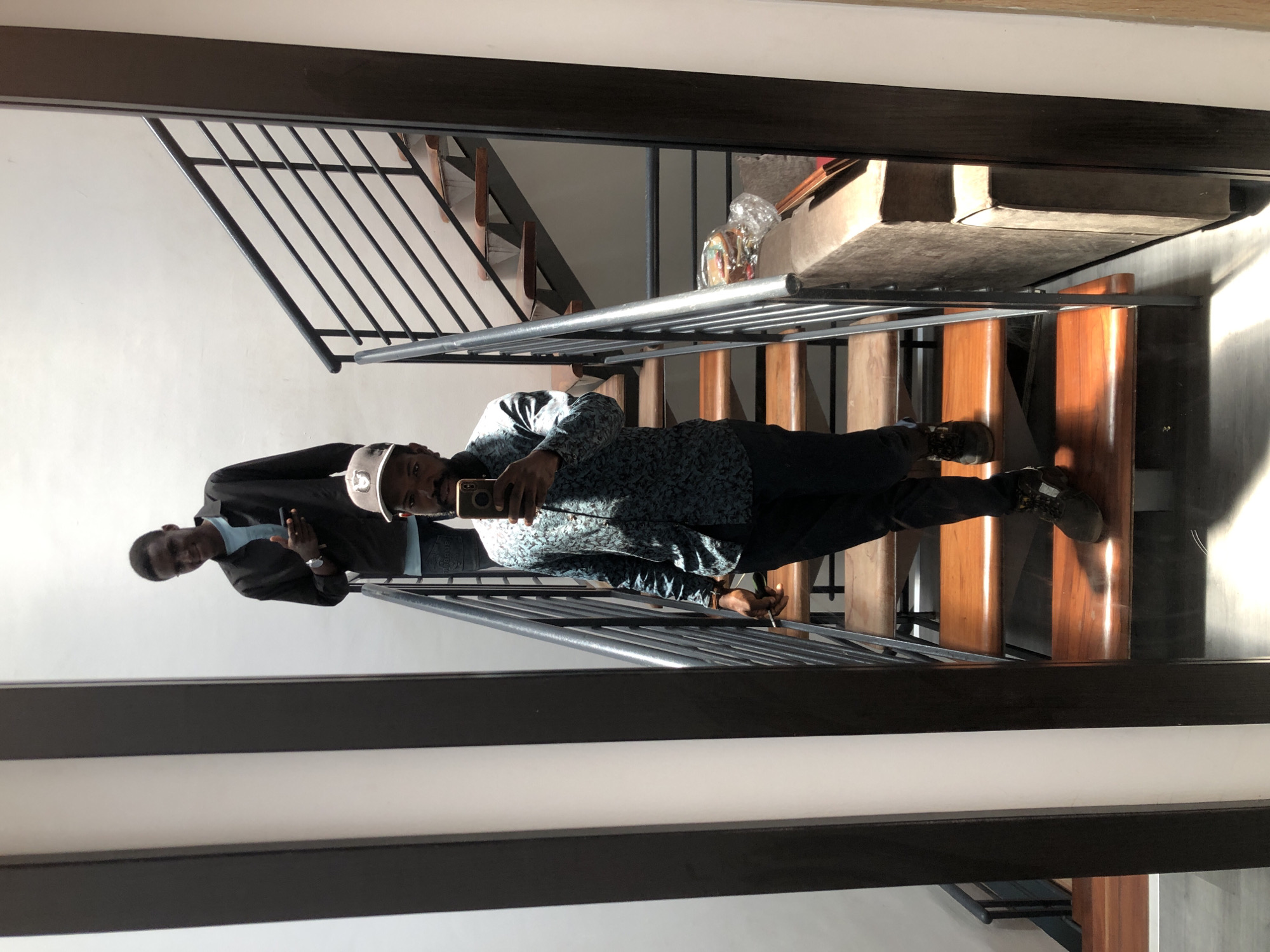 The image shows two young Black men on a modern wooden staircase with a metal railing. The man in the foreground is taking a mirror selfie with his phone, wearing a patterned blue shirt, dark pants, and grey sneakers. He is holding a cigarette in his right hand. A second man, wearing a black shirt and blue jeans, stands further up the stairs behind him and smiles at the camera, holding his phone. The staircase appears to be in an indoor, well-lit space, possibly a contemporary home or a commercial building. The background is a white wall. This scene is located in Igboefon, Nigeria.