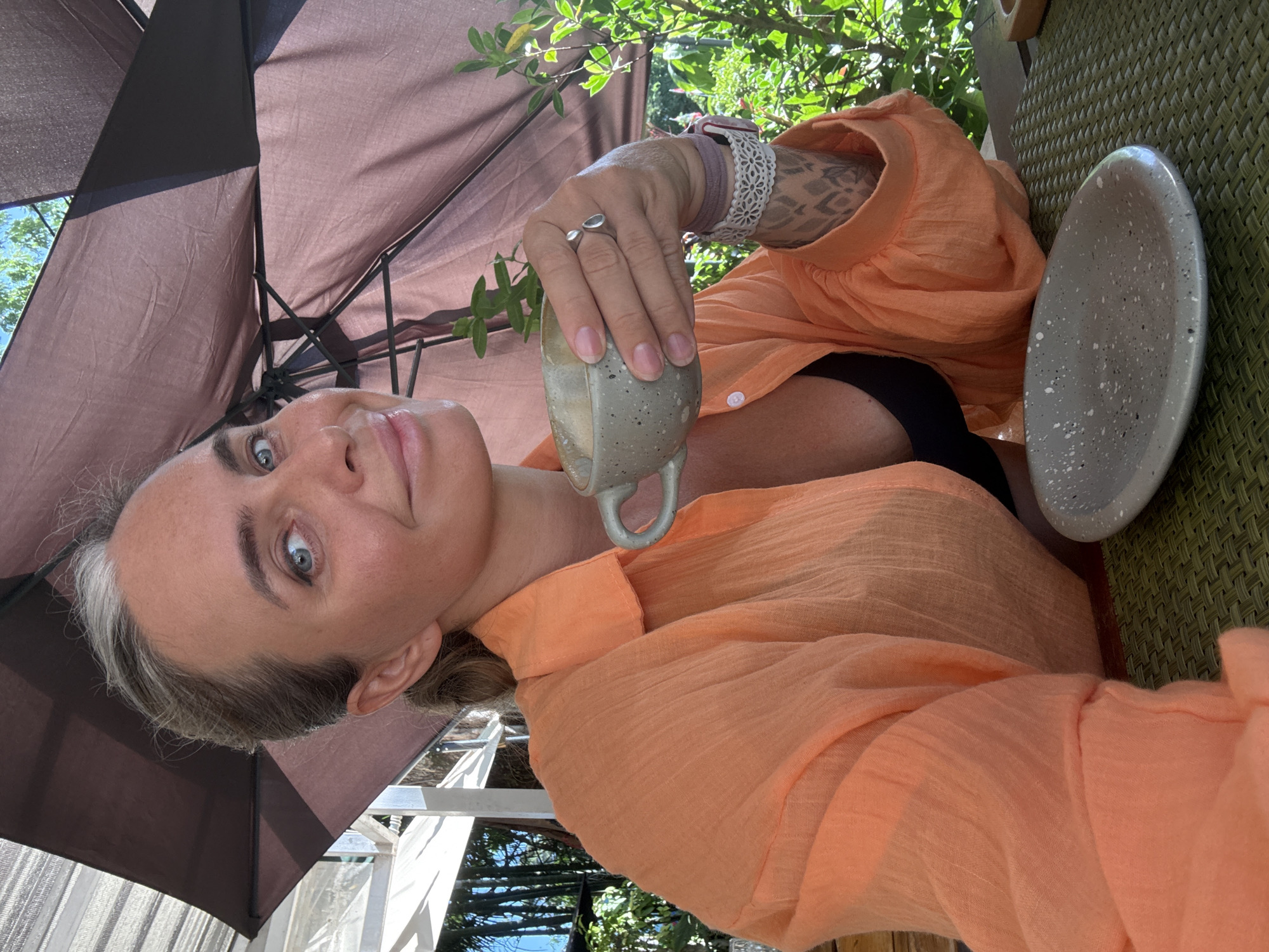 A medium shot captures a woman seated outdoors, holding a mug of coffee. She is wearing an orange, crinkled, long-sleeved button-up shirt and a black bra. Her left hand, adorned with a ring and a watch, cradles the mug, which contains a frothy, light brown beverage and a sprig of mint. A small, grey speckled plate rests on a textured green placemat in front of her. Sunlight filters through a brown umbrella, casting shadows on her face and shoulders. The background reveals lush green foliage and hints of white structures, suggesting an outdoor dining or seating area. The image appears to be taken on a sunny day in Ko Samui District, Thailand.