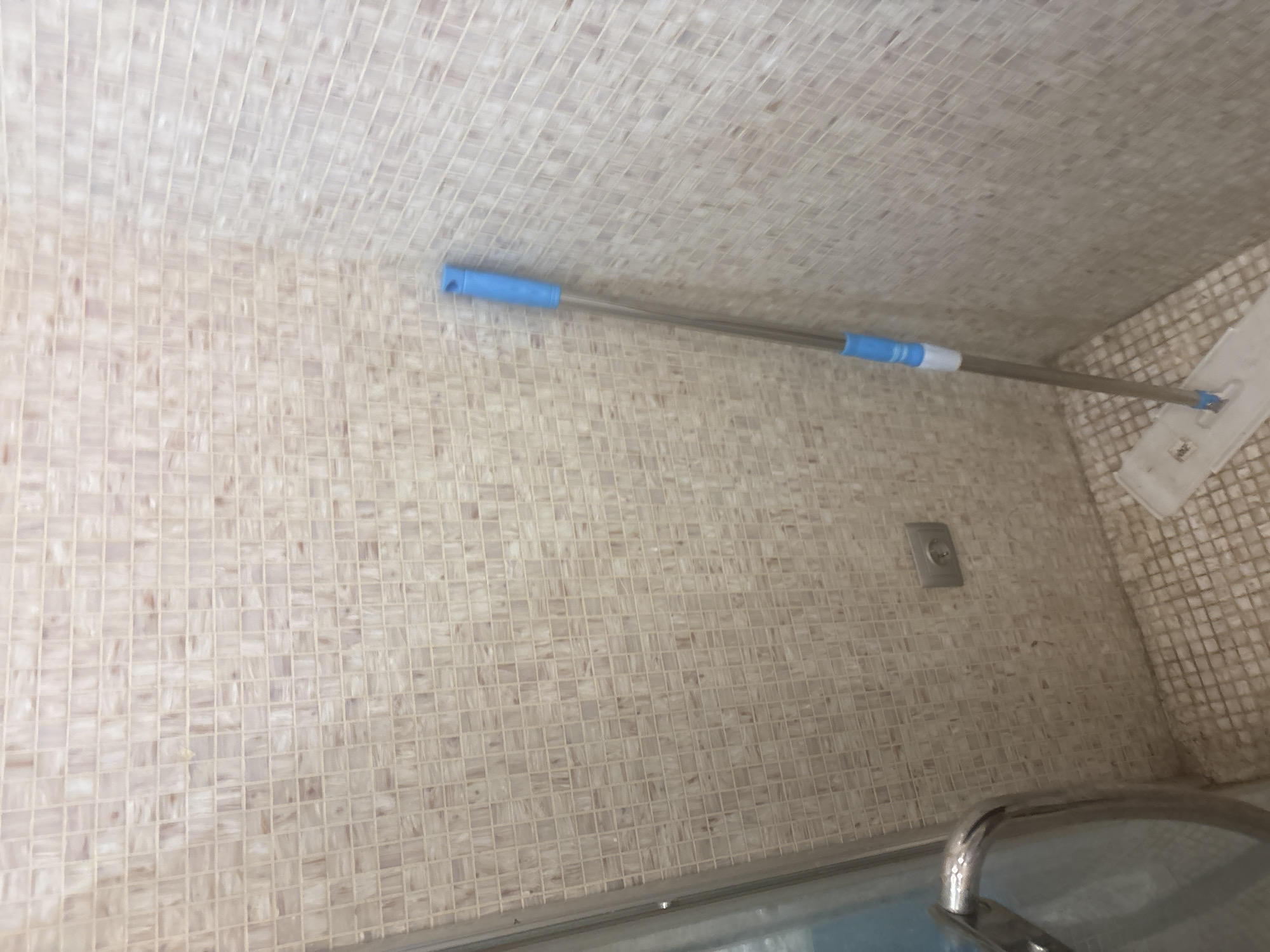 A mop with a blue handle stands in the corner of a bathroom. The walls and floor are covered in small, light-colored mosaic tiles. A chrome handle for a shower door is visible on the left side of the image. An electrical outlet is set into the wall. The scene is forensic and matter-of-fact, devoid of any contextual information about the city of Orekhovka, Russia.
