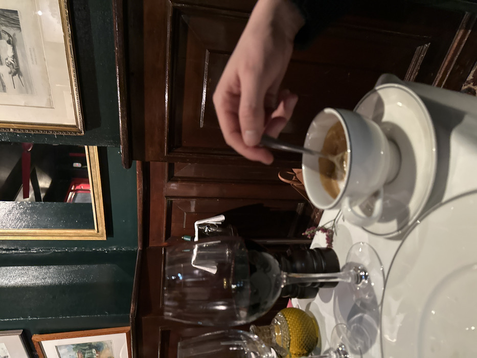 A hand stirs a white cup of coffee with a spoon. The cup rests on a saucer, part of a larger white plate setting. A lemon slice and two wine glasses occupy the foreground, slightly blurred. The background features dark wooden paneling. Framed artwork is visible on a green wall to the left.