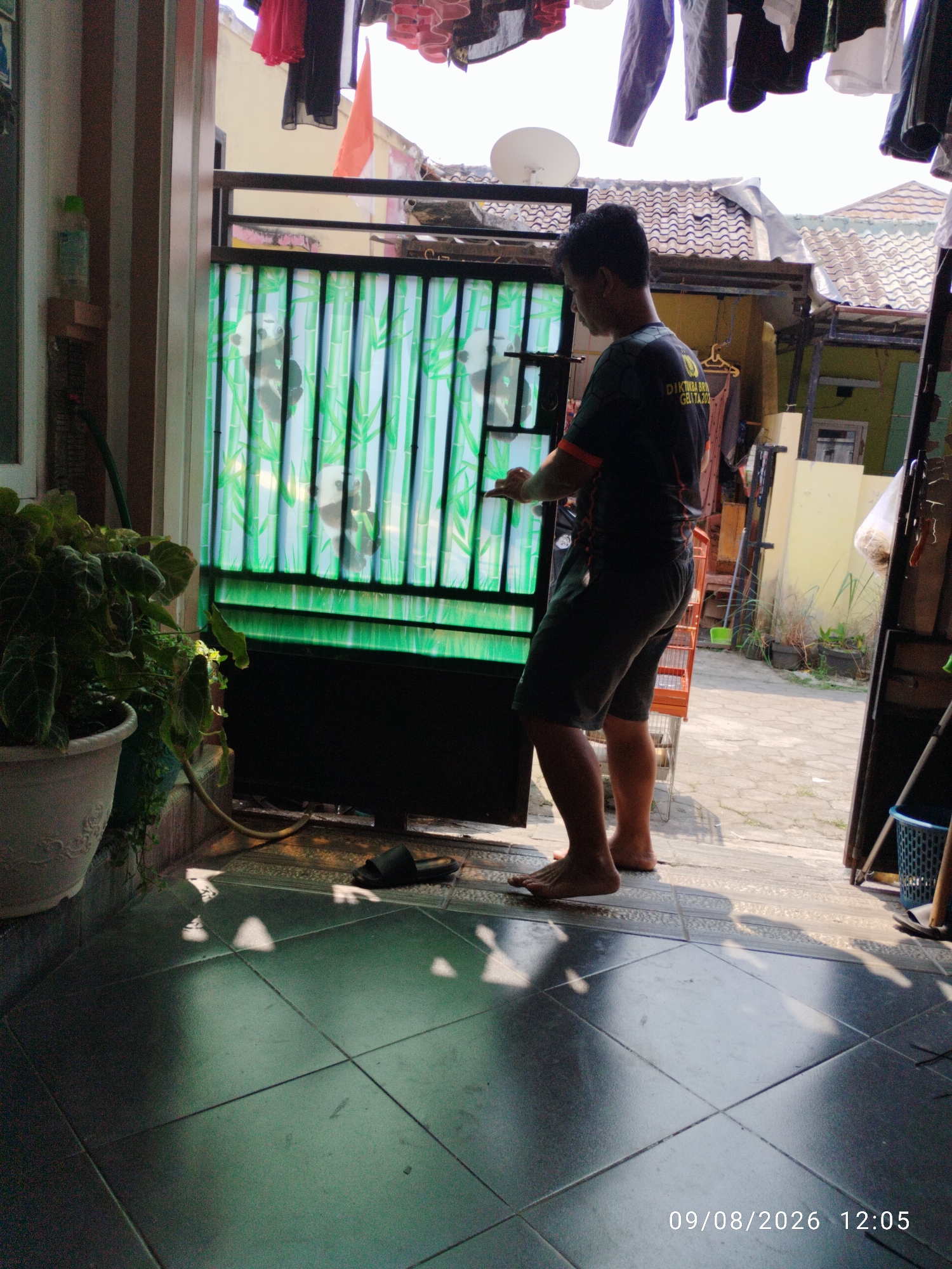 In a residential area of Bandar Lampung, Indonesia, a young man stands by a black metal gate, which is decorated with a green translucent material featuring a bamboo and panda motif. The man, wearing a dark t-shirt with an orange trim and dark shorts, is barefoot and appears to be interacting with the gate or something beyond it. His posture suggests he might be opening or closing it, or perhaps reaching for an object. To the left of the gate, a large potted plant with lush green leaves is visible. The ground is paved with dark square tiles, and sunlight casts distinct shadows. In the background, buildings with tiled roofs and a satellite dish are present, indicating a typical urban or suburban environment. Laundry is hanging on a line above the gate, suggesting domestic activity. The scene is dated 09/08/2026 at 12:05.