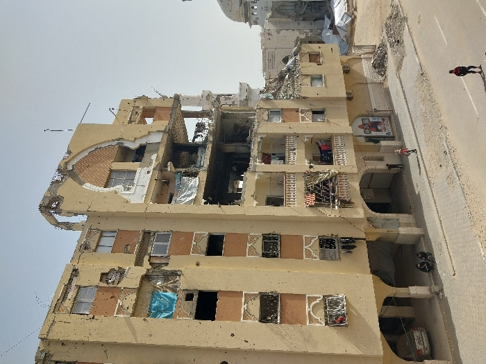 The image depicts a heavily damaged six-story building on a street in Khan Yunis, Palestinian Territory. The structure, primarily beige with orange accents on some walls, exhibits extensive destruction, including numerous small circular impact marks (bullet holes) across its facade and larger sections of collapsed walls and ceilings, particularly on the upper floors and the right side, exposing interior rooms. An arched architectural feature on the upper right side of the building is partially destroyed. Several windows are shattered or missing, with some openings covered by torn blue and white tarpaulins or sheets. Laundry is visible hanging on lines across multiple balconies, some with decorative balustrades, indicating recent or ongoing habitation despite the damage. The ground floor features an arched open-air area where a light-colored sedan is parked to the left.
On the street level in front of the building, two individuals are seated on a curb to the left, near the parked car. Further right, a small child in a dark outfit and an adult in a light red top and dark pants walk on the sidewalk. A male figure in a red and black tracksuit walks alone in the street's foreground, moving right. The street itself is paved but shows signs of wear and loose debris. To the right of the main building, debris piles are visible on the ground. In the background, further to the right, another large structure with a prominent dome and classical architectural elements, heavily damaged, is visible; this structure is identifiable as the Great Mosque of Khan Yunis. The sky is clear and light blue.