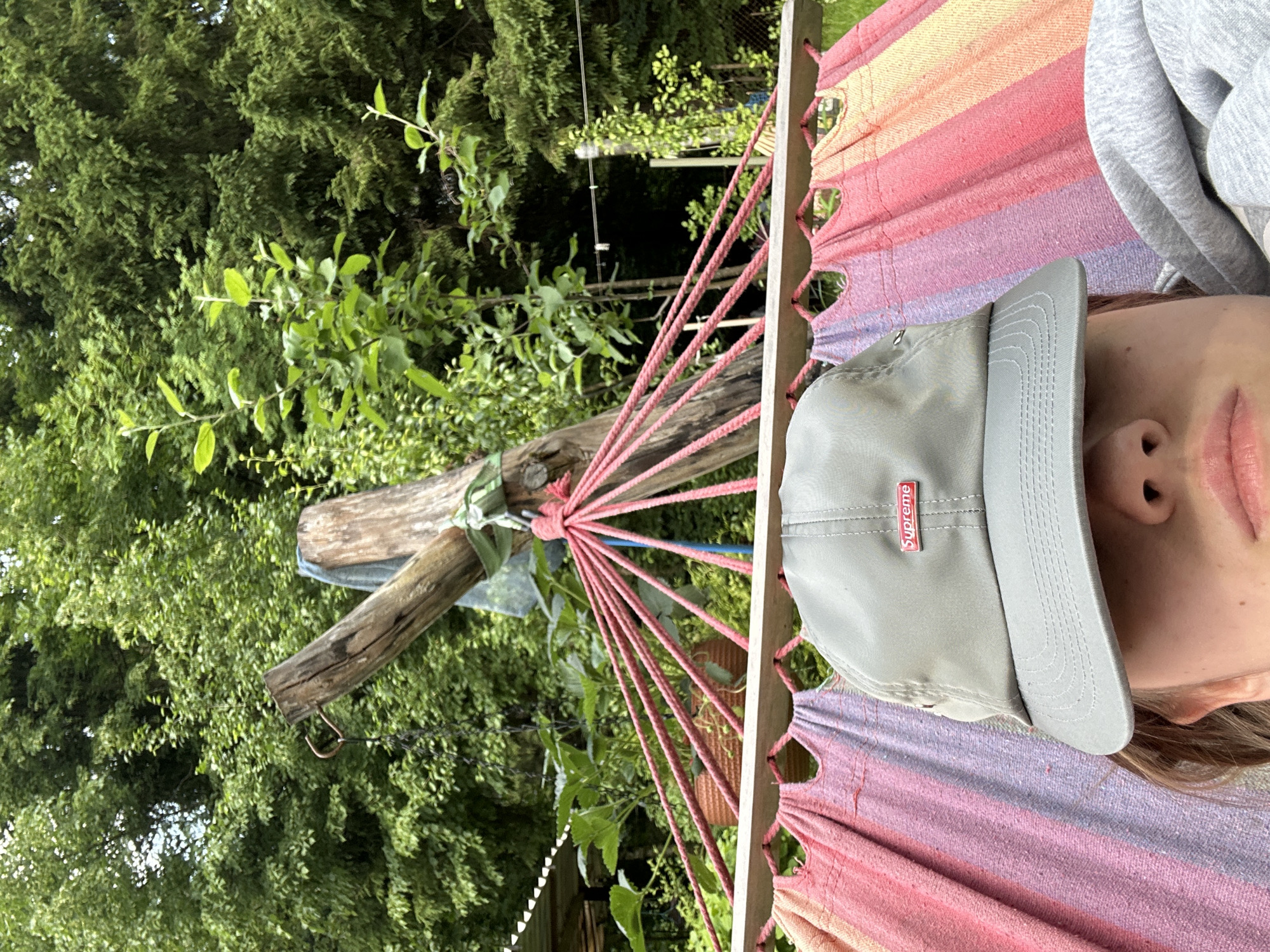The image shows a close-up of a person wearing a gray baseball cap with a red "Supreme" logo. The person is lying in a hammock with a colorful striped fabric. The background features lush green trees and vegetation, suggesting an outdoor setting. There are wooden posts and ropes visible, part of the hammock's structure. The overall scene conveys a relaxed, summery atmosphere. The location is not specified in the image itself.