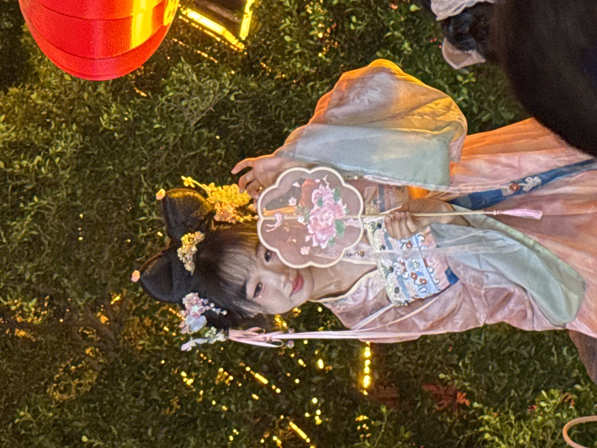 A young East Asian woman in traditional Chinese attire stands outdoors at night. She wears a pink and light blue robe adorned with decorative trim. Her hair is styled in elaborate buns with floral accessories. She holds a small, decorative fan with a floral design in front of her face. The background is a dense green bush illuminated by numerous small, yellow lights and a large red lantern. A portion of another person's head and shoulders is visible in the foreground, obscuring the lower right of the frame. The scene appears to be in Yuzhong District, China, possibly during a festival or cultural event.