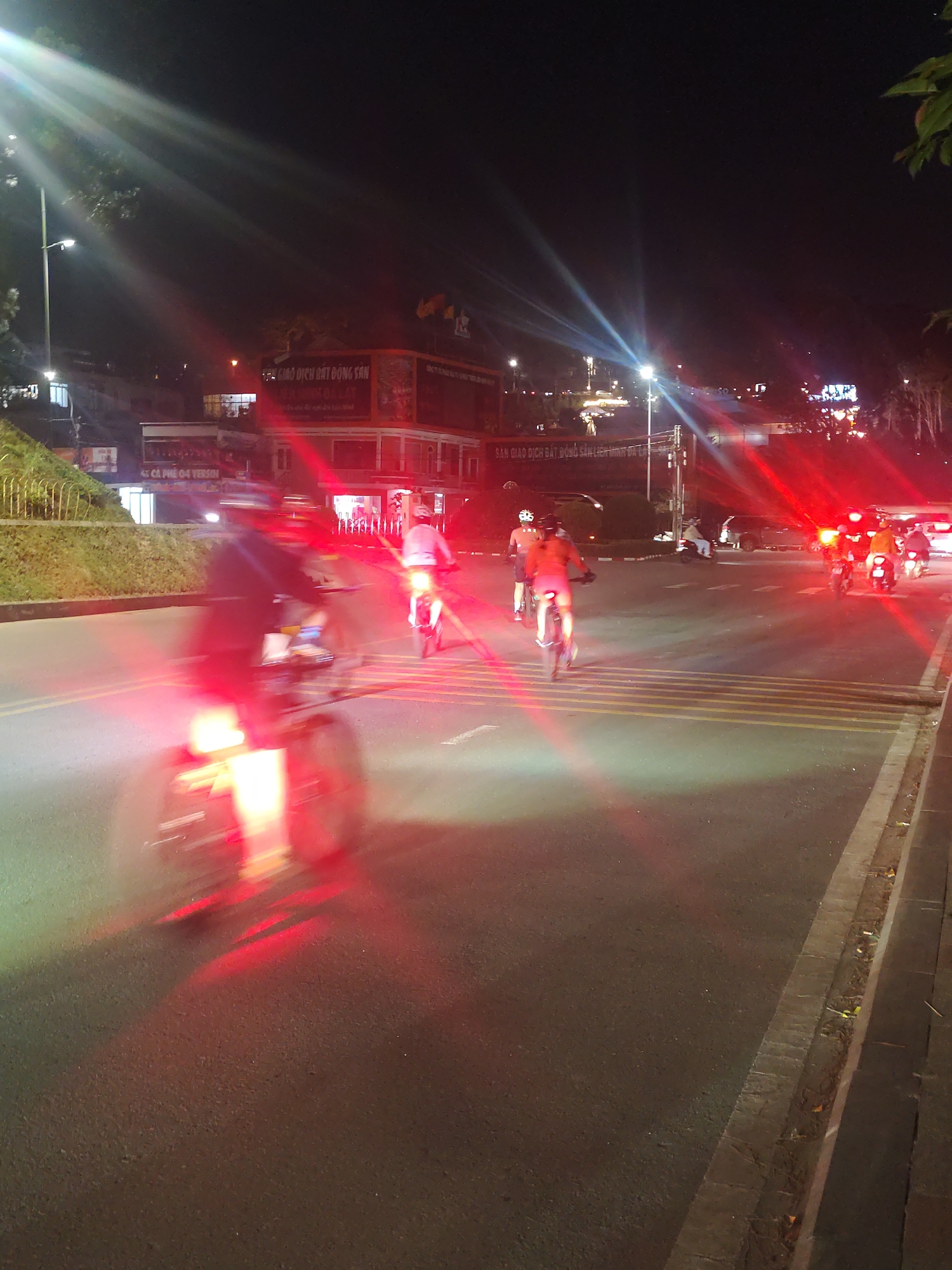 The image depicts a night scene on a city street, likely in Da Lat, Vietnam, given the contextual clues from the text on buildings and the overall ambiance. Several individuals are riding bicycles and motorcycles, with their headlights and taillights creating streaks of light due to the motion blur and the low-light conditions.
In the foreground, a person on a bicycle is prominently visible, their red taillight creating a strong glow on the asphalt. The wheels of the bicycle are blurred, indicating movement. To the right and further down the street, other individuals on bicycles and motorcycles are also captured in motion. One person on a bicycle in the mid-ground is wearing a light-colored shirt and dark shorts. Another individual on a motorcycle behind them is also illuminated by their vehicle's lights.
Further in the background, there are buildings with illuminated signage. One sign, partially visible, appears to read "SÀN GIAO DỊCH BẤT ĐỘNG SẢN", which translates to "Real Estate Exchange". Another sign reads "45 CÀ PHÊ 04 VERSIN". The street has clear yellow markings indicating lanes. Trees and vegetation are visible on the left side of the street, and a sidewalk with some debris is on the right. The sky is dark, consistent with a nighttime setting, with some light sources creating lens flare effects. The overall atmosphere suggests a busy urban environment at night, possibly a popular area for evening cycling or commuting.