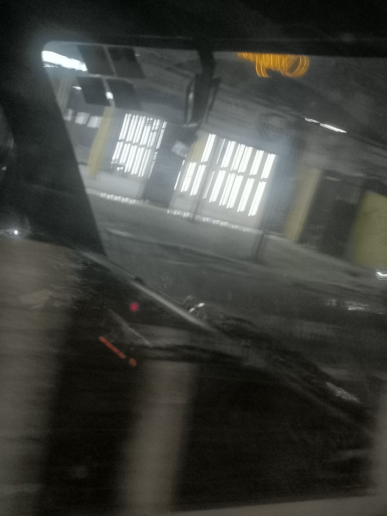 The image, taken from inside a vehicle at eye level, appears to be of a building with large windows or openings that are illuminated by fluorescent lights. The building structure suggests it might be a warehouse, industrial facility, or perhaps a large parking garage, possibly located in Depok, Indonesia. The perspective is slightly angled, and there is a reflection on the windshield, possibly from the interior of the vehicle or from the lights. A windshield wiper is visible in the foreground. There are some blurry streaks of orange and red light, which could be reflections or distant traffic signals. The overall lighting is dim and the image quality is somewhat poor, with a grainy texture and a lack of sharp focus, suggesting it was taken in low light conditions or with a low-quality camera.