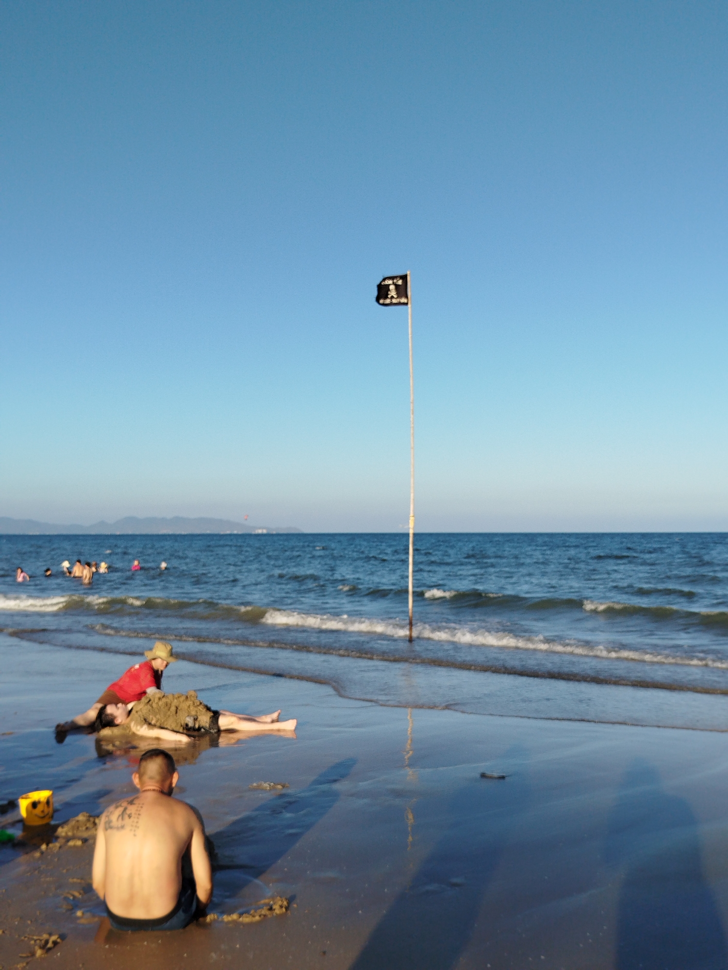 The photograph was taken at a beach in Ho Chi Minh City, Vietnam, with a clear blue sky and calm blue water. A black pirate flag is flying on a tall pole in the shallow water. In the foreground, a shirtless man sits in the sand with his back to the camera, revealing a tattoo on his back. Nearby, a yellow bucket sits in the sand. Further up the beach, a person in a red shirt and a straw hat is covering another person lying on the ground with sand. Several people are swimming in the sea in the distance. The wet sand reflects the sky and the shadows of the photographer.