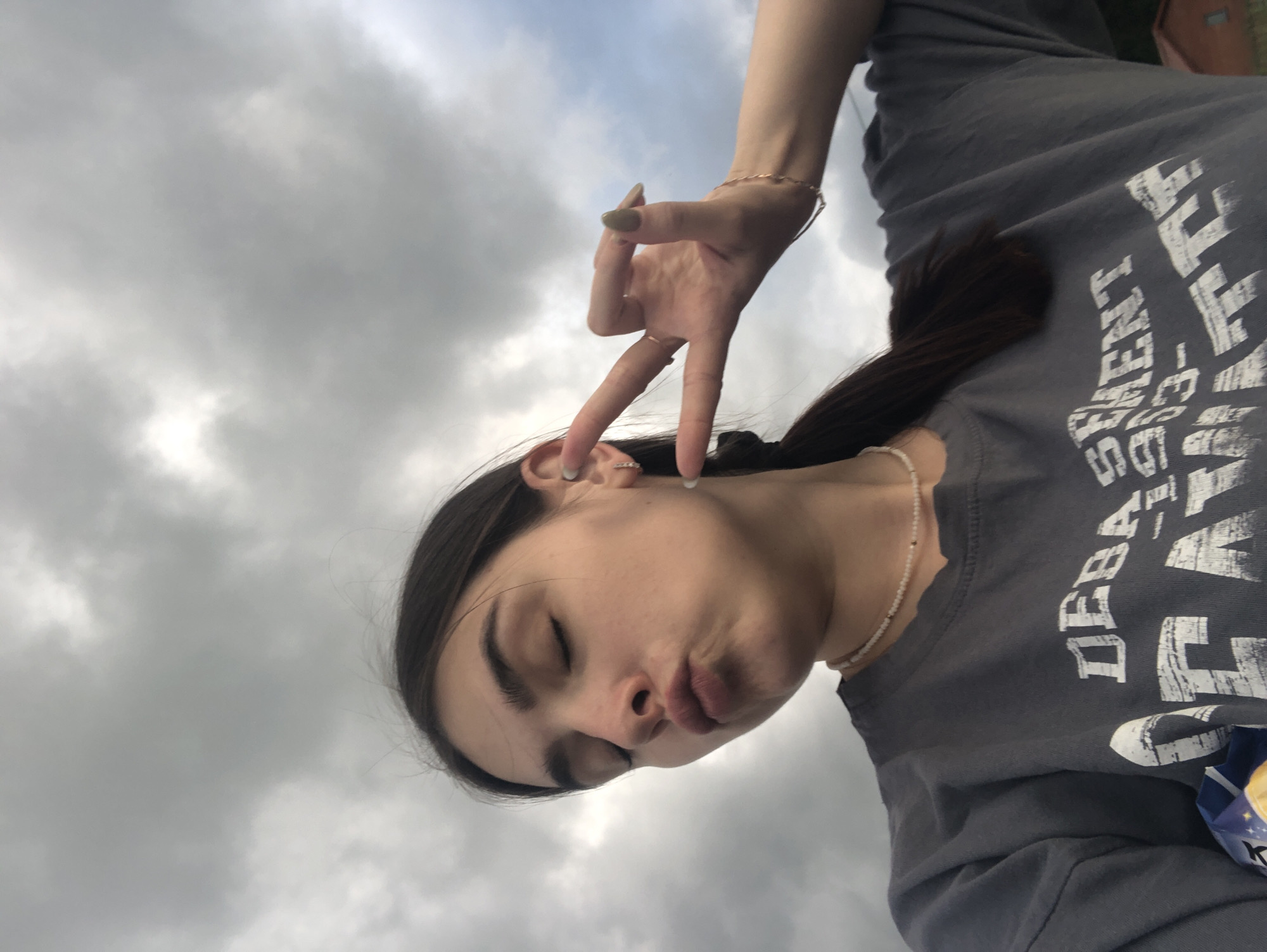 This media file features a close-up, rotated shot of a young woman posing against a cloudy sky. She is wearing a dark gray t-shirt with white text that appears to be a distressed graphic print. On her neck, she wears a delicate beaded necklace. Her left hand is raised, with two fingers extended in a peace sign gesture, adorned with rings on her middle and index fingers, and a bracelet on her wrist. Her eyes are closed, and her lips are puckered, giving her a playful or perhaps contemplative expression. The lighting suggests it might be daytime, given the brightness in the sky, but the heavy cloud cover indicates overcast weather. The overall mood is casual and lighthearted. There is visible text on the t-shirt, but it is partially obscured and difficult to read in its entirety. The background is dominated by a dense, gray cloud formation, with hints of lighter sky peeking through. There are no clear geographical cues to definitively place the location within Noviy, Russia, other than the provided context.