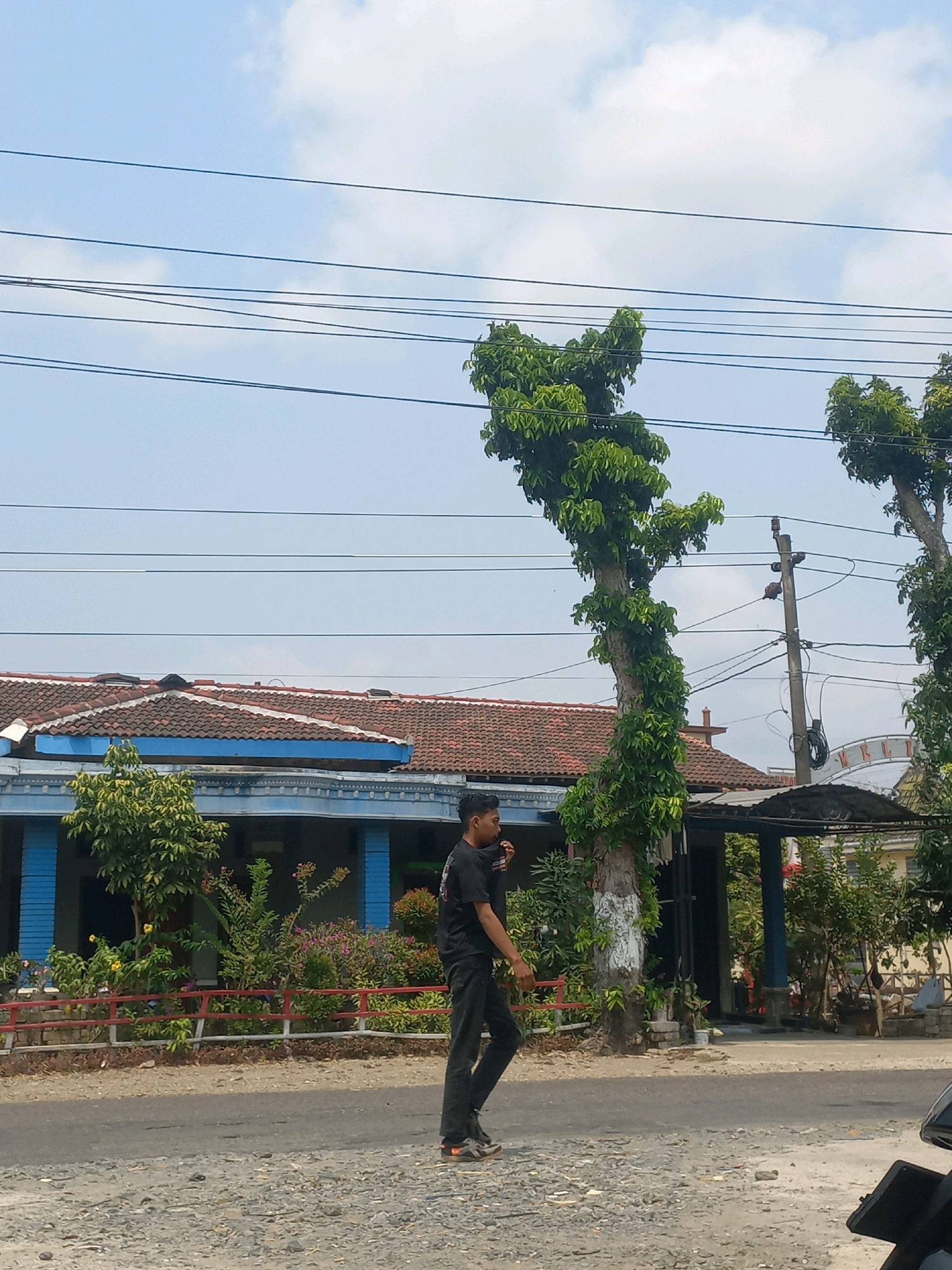 In Baturetno, Indonesia, under a partly cloudy sky, a young man in a black t-shirt and black pants walks across a gravelly road. He is positioned in the left-center of the frame, facing left and looking down. In the background, a building with a red tiled roof and blue trim is visible. Several trees with green foliage are situated in front of the building, and power lines stretch across the sky above. A utility pole with electrical equipment stands to the right of the road.