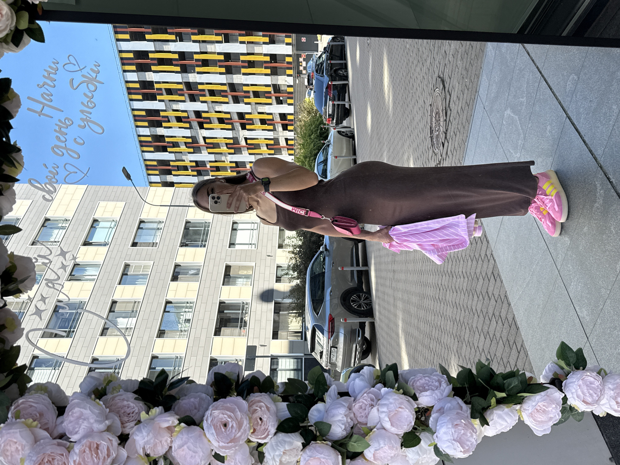 A full-body mirror selfie shows a woman with long brown hair, wearing a long brown spaghetti-strap dress, a pink crossbody bag with a white strap, and bright pink sneakers with yellow stripes. She is holding a pink and white striped garment. The background, reflected in the mirror, displays a modern building with a facade of alternating beige and pale yellow rectangular panels, and a multi-story parking garage with a vibrant facade of vertical yellow, red, and black rectangular sections. To the left of the reflection, in the foreground, is a large, ornate floral arrangement of light pink and white peonies. The setting appears to be a street or public walkway in Saint Petersburg, Russia, with paved ground and parked cars visible in the background. The text "Начни свой день с улыбки" ("Start your day with a smile") is written in white cursive script across the top of the mirror.