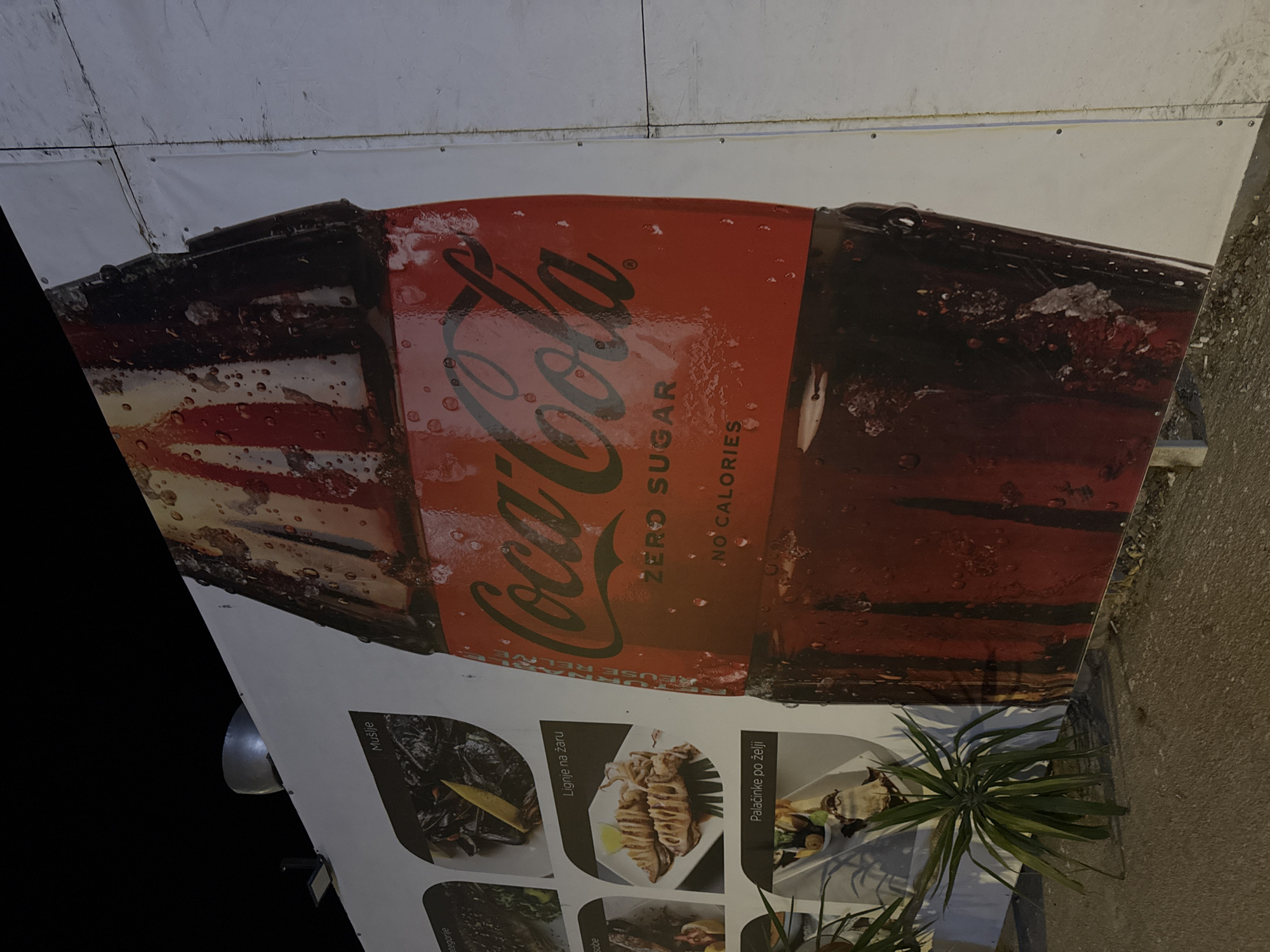 A large, illuminated banner featuring a Coca-Cola Zero Sugar bottle dominates the scene. The banner is affixed to a temporary wall constructed from white panels, likely part of an outdoor setup. To the left of the Coca-Cola advertisement, a series of smaller images display food items, including what appears to be calamari and mussels, with accompanying text in a language that appears to be Cyrillic or Latin alphabet. A potted plant with long, green leaves is situated in the foreground, casting a shadow on the concrete ground. The environment suggests an outdoor, possibly commercial setting at night, with the dark sky visible above the banner. The overall impression is a brightly lit advertisement juxtaposed with food offerings. The location is identified as Budva, Montenegro.