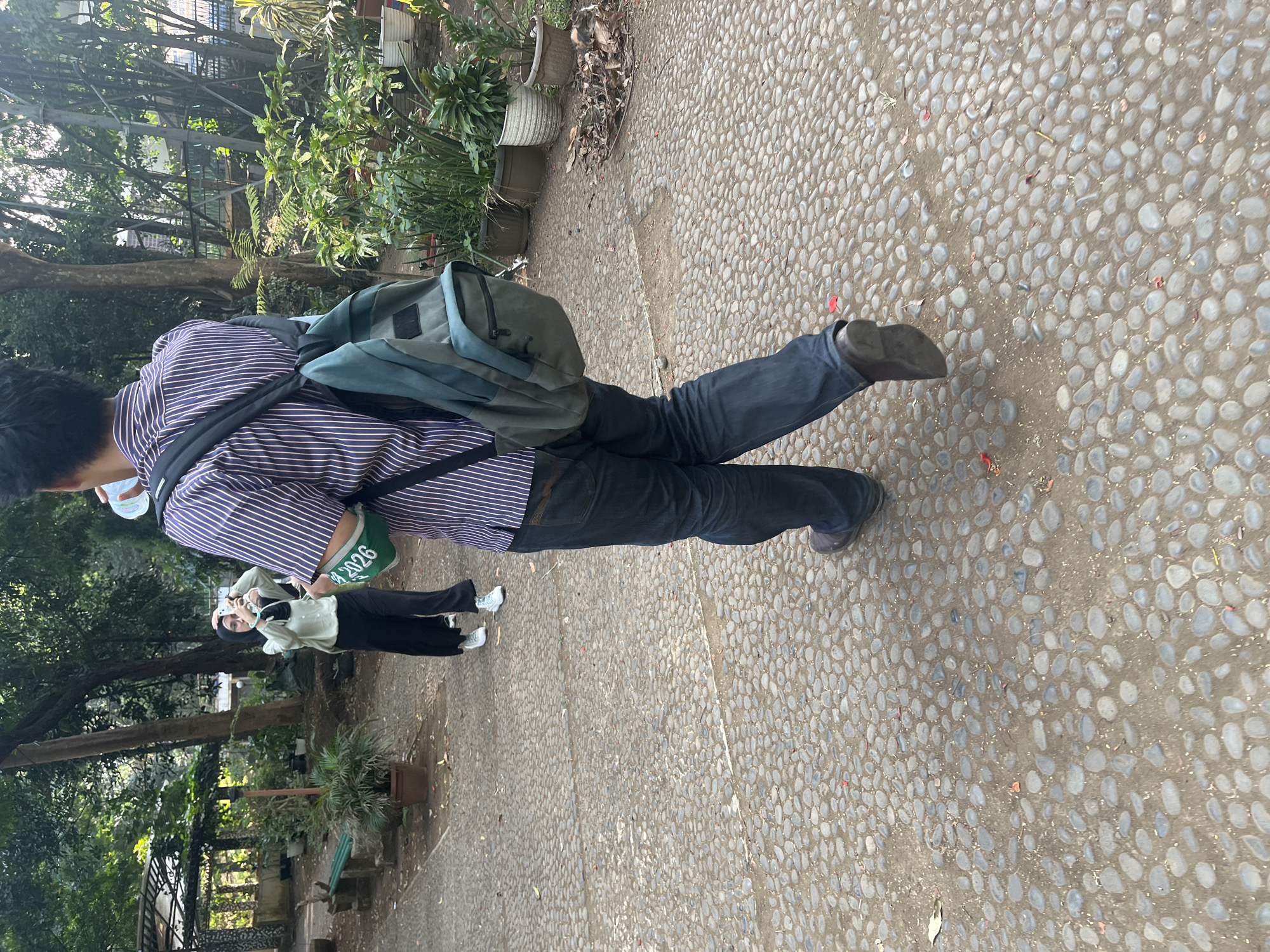 The image depicts a paved outdoor area, likely a park or garden, with a cobblestone-like ground surface. Two individuals are visible. In the foreground, a person is walking away from the camera, wearing a striped shirt, dark jeans, and a backpack. They are holding a water bottle to their mouth. A green armband with the text "2026" is visible on their left arm. In the background, a second person, wearing a headscarf, light-colored shirt, and dark pants, is taking a photo with a smartphone. The environment is characterized by lush green trees and potted plants. The scene appears to be set in a public outdoor space.