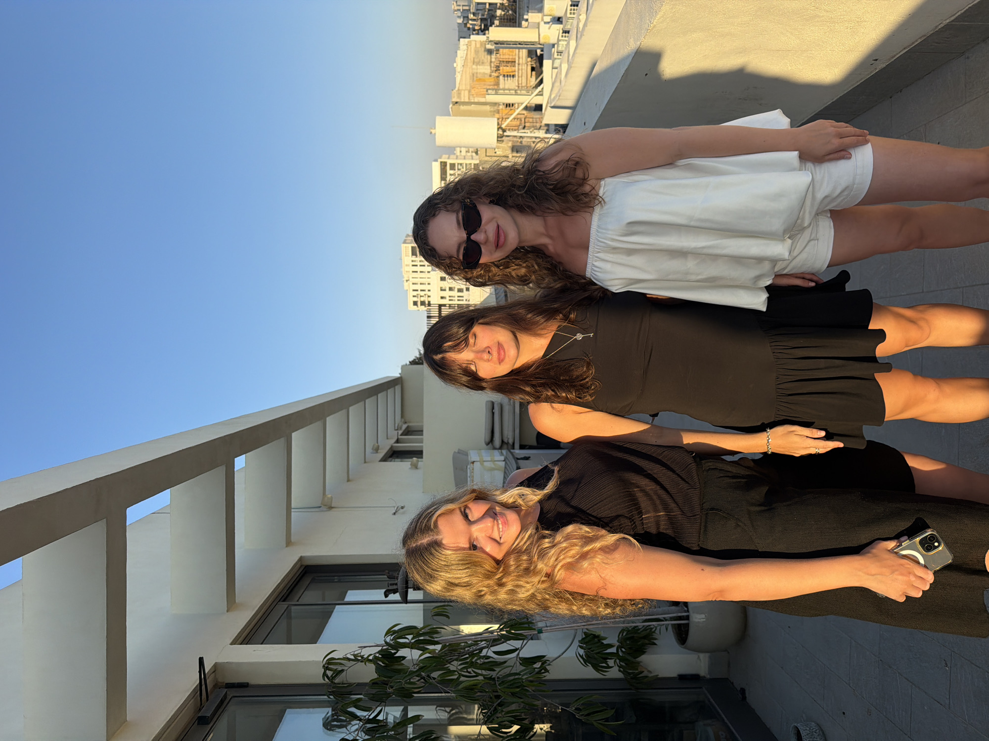 Three young women stand on a balcony against a backdrop of Tel-Aviv buildings under a clear blue sky. The woman on the left has long blonde curly hair and wears a black sleeveless top and a black skirt. She holds a phone in her left hand. The woman in the middle has long dark hair and wears a black dress with a ruffled hem. The woman on the right has curly brown hair and wears sunglasses, a white strapless top, and white shorts. The balcony features a white railing and a building with modern architectural elements. A potted plant stands to the left of the women. The setting suggests a rooftop gathering or vantage point overlooking the city.