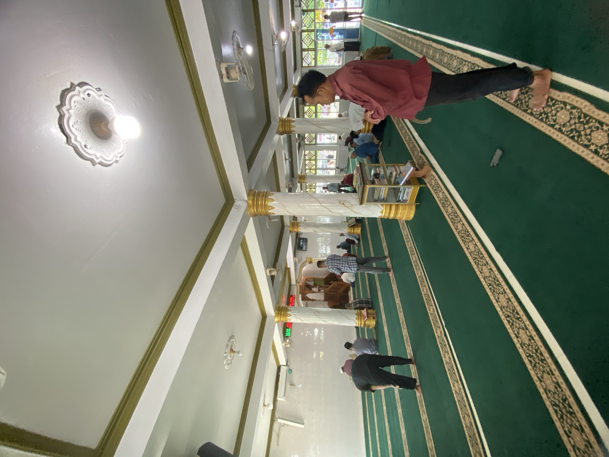 The image captures the interior of a mosque in Lhokseumawe, Indonesia, likely during the daytime. The perspective is from inside the main prayer hall, looking towards the front of the mosque. The floor is covered with a vibrant green carpet, adorned with ornate gold and cream patterns, and marked with white stripes delineating prayer rows.
Several men are present, engaged in prayer or contemplation. In the foreground, a man wearing a red long-sleeved shirt and black pants stands barefoot on the carpet, facing towards the mihrab. A white mobile phone is placed on the carpet near his feet. Further into the hall, other men are visible in various stages of prayer, including standing and prostrating. They are dressed in casual clothing, with some wearing traditional prayer caps.
The mosque's architecture features tall, white, marble-patterned pillars accented with decorative golden bases and capitals. The ceiling is white with prominent golden trim, fitted with ornate white medallions from which simple light bulbs illuminate the space. Small ceiling fans are also mounted. A red digital display on one of the pillars shows '06:05', possibly indicating the time. In the background, a more elaborate wooden structure, potentially a mihrab or minbar, is discernible. Windows along one wall allow natural light to filter in, complemented by artificial lighting. A small, golden-hued bookshelf or stand filled with books is situated near a pillar. A white wall-mounted air conditioning unit is visible on the left wall.
The scene conveys a calm and reverent atmosphere, typical of a place of worship. The bright lighting, a mix of natural and artificial sources, enhances the sense of tranquility within the mosque.