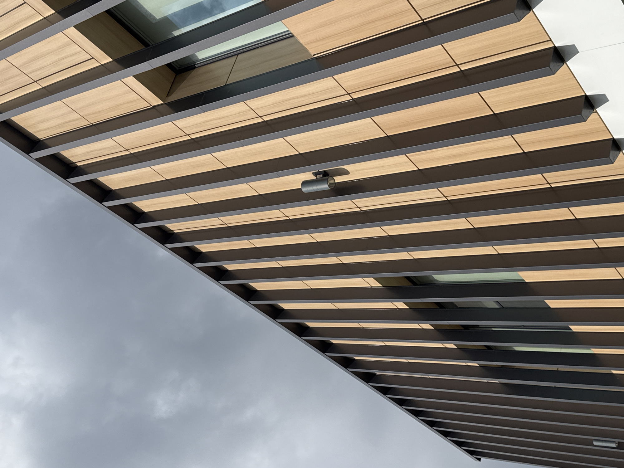 The image shows an exterior view of a modern building's architectural design, looking upwards. The ceiling is constructed with horizontal wooden slats interspersed with dark, linear elements, likely metal beams or supports. These slats are arranged in a repeating pattern, creating a sense of depth and rhythm. Several rectangular glass windows are visible between the wooden panels, reflecting the sky. A small, cylindrical light fixture with a metallic finish is attached to one of the beams. The building's exterior contrasts with a cloudy, overcast sky, suggesting either dawn, dusk, or a generally gloomy weather condition. The lighting indicates diffused daylight. No people are visible in the image. The scene appears to be an urban or suburban setting, focused on architectural detail rather than a specific event. The overall impression is one of modern, clean design. There is no visible text in the image.