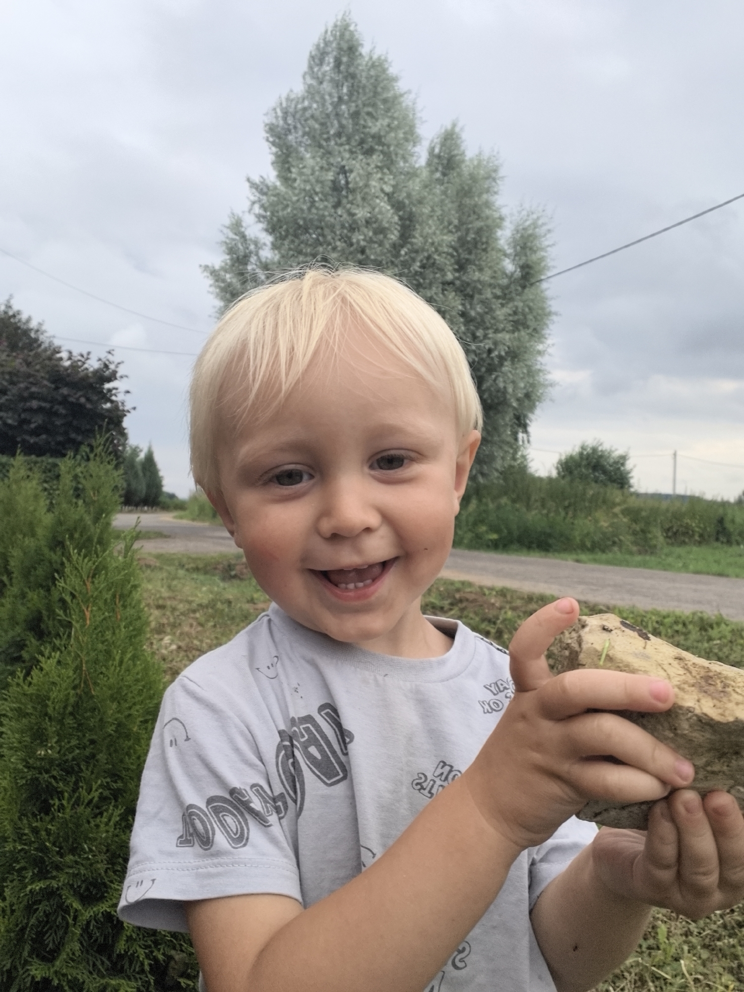 A young, fair-skinned boy with blond hair is holding a rock in an outdoor setting with an overcast sky. He is wearing a gray t-shirt with a repeating smiley face pattern and dark gray text. The boy is smiling with his mouth open, revealing teeth. He is positioned in the foreground, with a blurred background that includes trees, grass, and a road. To the left of the boy is a small, green, conical bush. The boy is looking towards the camera and holding the rock with both hands, showing his right hand more prominently. The rock is light brown and appears to be irregular in shape with some plant matter attached.