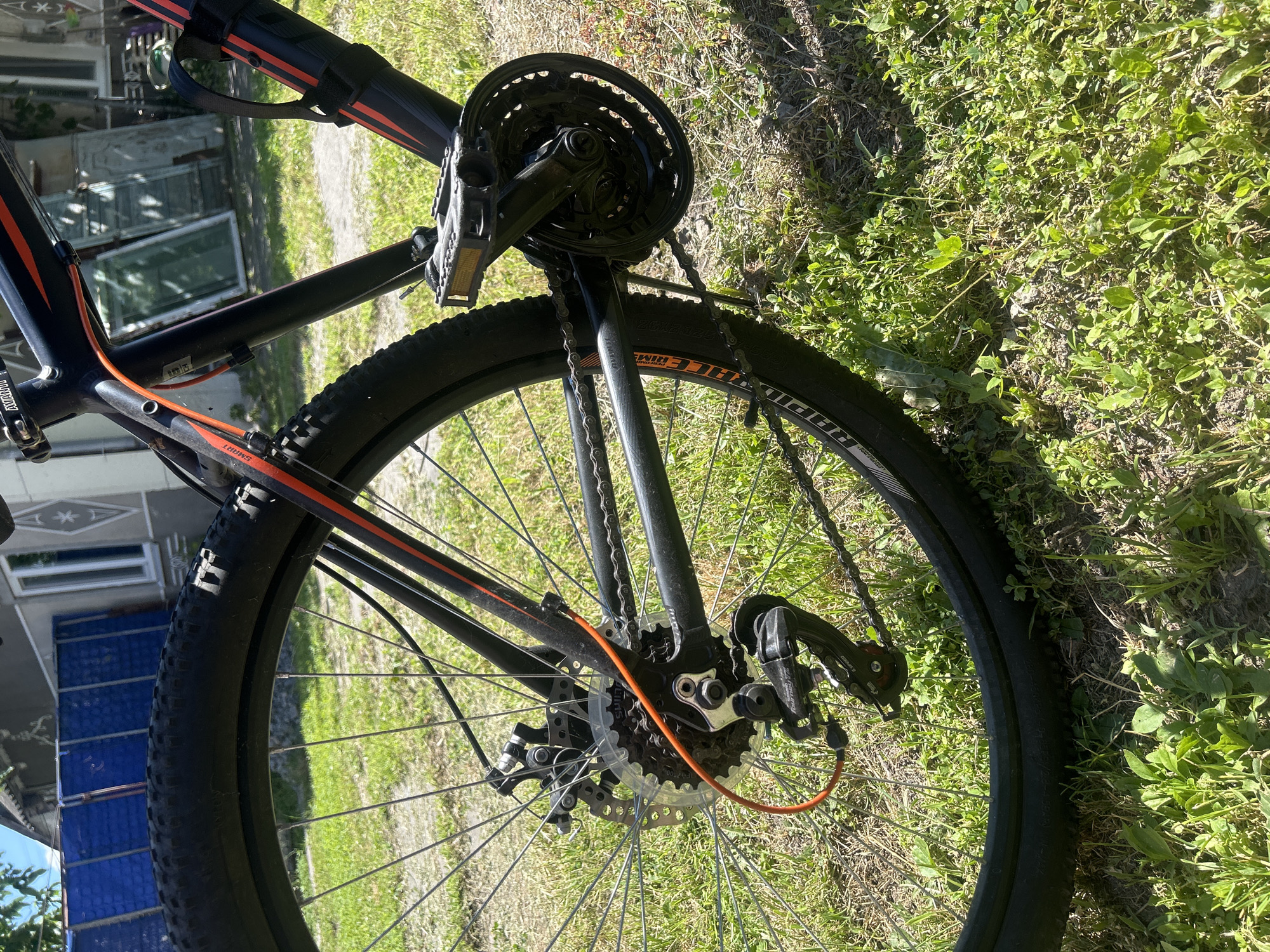 The image shows a close-up, low-angle view of the rear wheel and drivetrain of a black bicycle parked on a grassy area. The bicycle has orange accents on the frame and orange cable housing. The tire has "RAPID" written on it. In the background, a partially visible house with a blue fence and some stacked items are present. The environment suggests a residential yard on a sunny day. This scene is located in the city of Bila Tserkva, Ukraine.