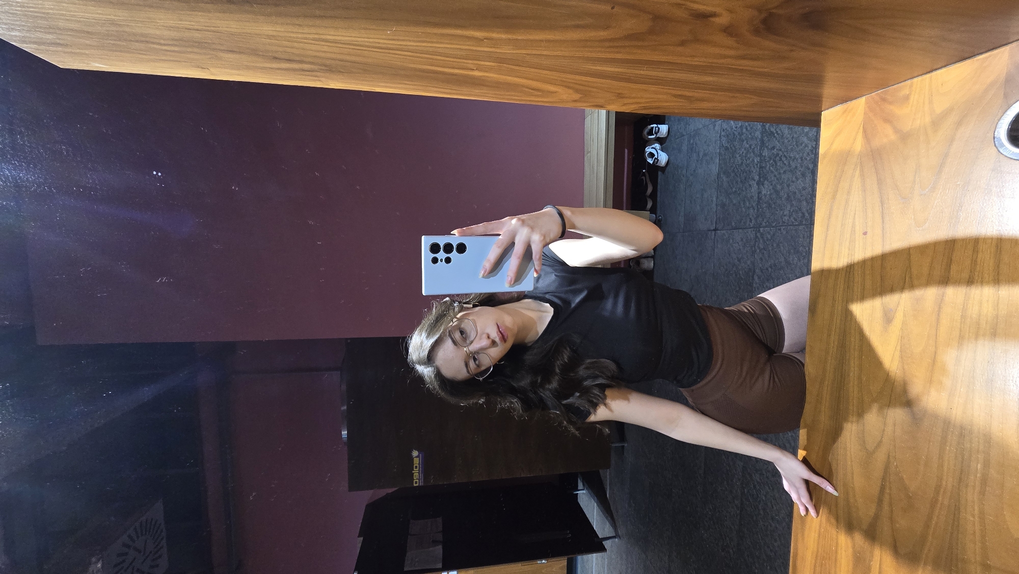 A woman is taking a selfie in a mirror. She is wearing a black top and brown shorts. She has long brown hair and is wearing glasses. The background includes a maroon wall, a black locker, and a wooden countertop. There are shoes on a shelf near the wall. The photograph was taken in Stara Zagora, Bulgaria.