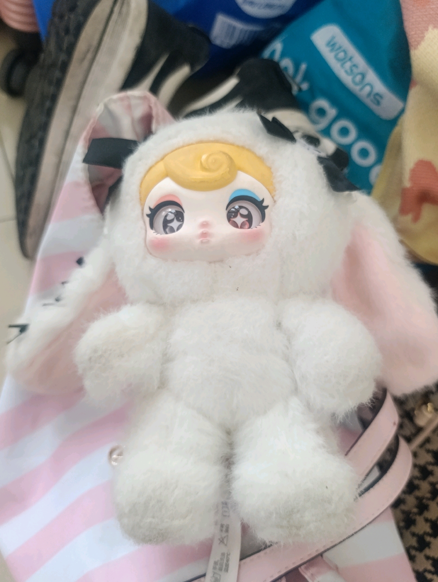 The image shows a close-up of a white, fluffy stuffed toy resembling a bunny, with a doll-like face. The toy has large, detailed eyes and pink blush on its cheeks. It is positioned on top of a pink and white striped bag. In the background, parts of a black and white checkered fabric and a blue bag with the "Watsons" logo are visible, along with the sole of a shoe. The scene appears to be indoors, possibly in a home environment. There is no indication of the location being Cihanjuang, Indonesia.