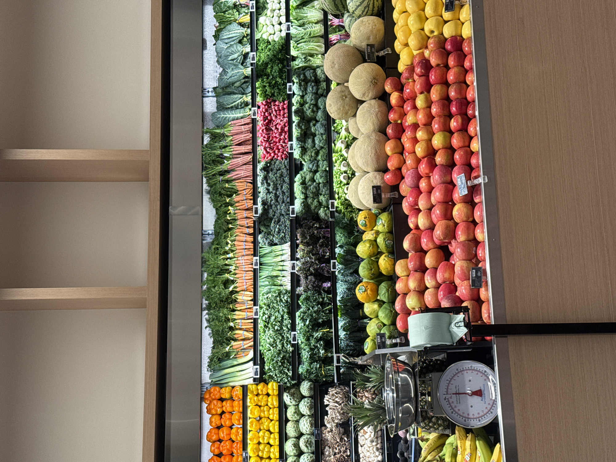 This image shows a produce section in a grocery store, likely located in West Hollywood, California, United States of America. The produce is neatly arranged in vertically stacked rows. The rows consist of various fruits and vegetables, including collard greens, lettuce, watermelon, cantaloupe, yellow apples, red apples, carrots, radishes, kale, broccoli, papayas, oranges, yellow bell peppers, green artichokes, mushrooms, pineapples, bananas, and possibly other items. In the lower right corner is a scale, with a green produce bag sitting nearby. The grocery section is well-lit. To the left of the produce section is a set of empty shelves. To the right, is a wall.