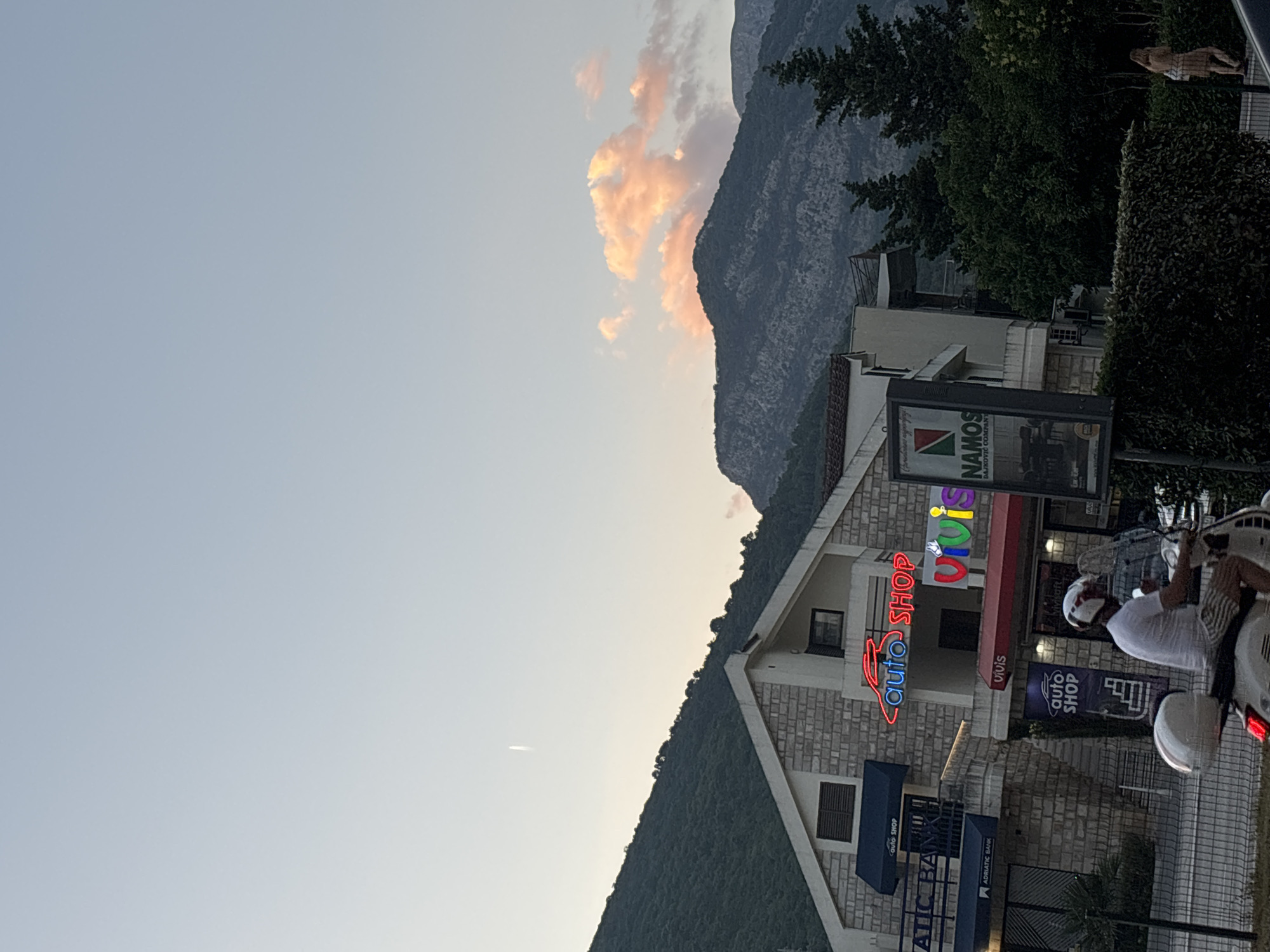The image captures a street scene in Budva, Montenegro, during the late afternoon or early evening. In the foreground, a person on a white Vespa scooter is seen from behind, wearing a white t-shirt, striped shorts, and a white helmet. They are riding towards the right side of the frame, on a paved road with a metal fence bordering the left.
Behind the road, a multi-story building with a stone facade features several businesses. A prominent neon sign in red and blue reads "auto shop." Another sign in bright, multi-colored neon letters spells out "VIVIS." An "auto SHOP" sign in blue is also visible on a dark blue awning. Below the "auto shop" neon sign, a red awning covers a store. To the left of these businesses, an "ADRIATIC BANK" sign is displayed on a dark blue awning. A large billboard for "NAMOS BAJKOVIC COMPANY" is positioned on the right side of the building.
The background consists of lush green mountains that are partially obscured by trees. The sky above is a pale blue, transitioning to a lighter hue near the horizon, with scattered clouds tinged with pink from the setting sun. A thin, white contrail from an aircraft is visible high in the sky. The overall atmosphere suggests a warm, pleasant evening in a coastal town.