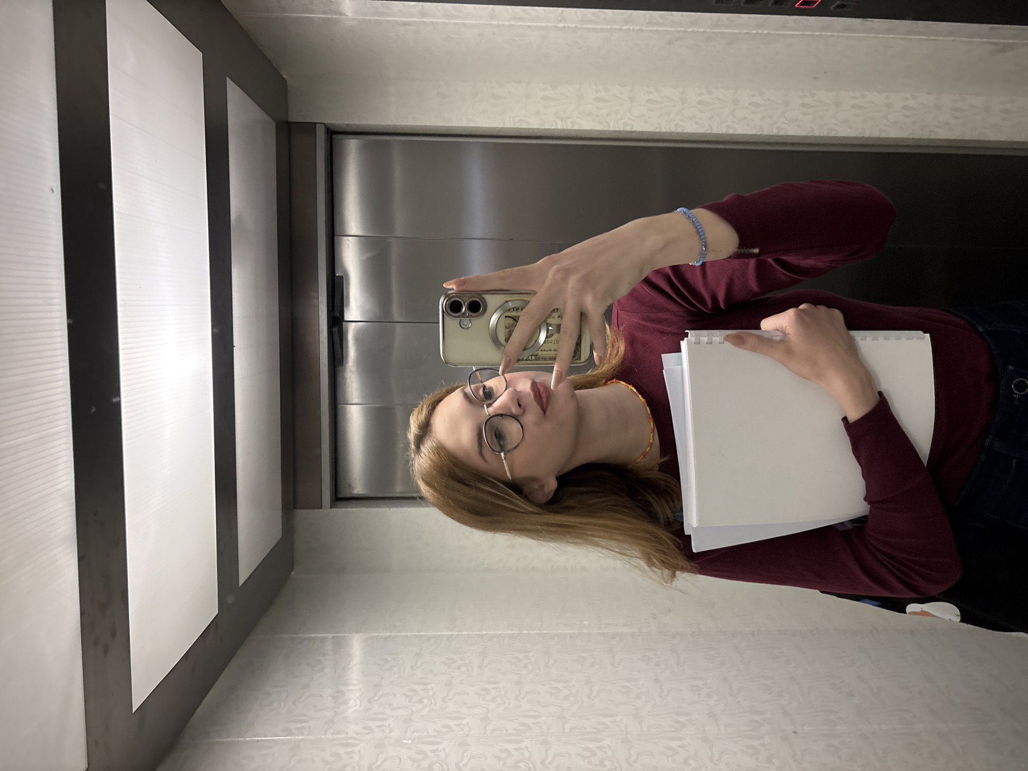 A young woman takes a selfie in an elevator. She wears glasses, a maroon long-sleeved shirt, and blue jeans. She holds a notebook in one hand and a cell phone in the other, capturing the image in the elevator mirror. The elevator walls are made of stainless steel and white panels. A red digital display is visible above the mirror. The location of Kragujevac, Serbia, cannot be determined from the image.