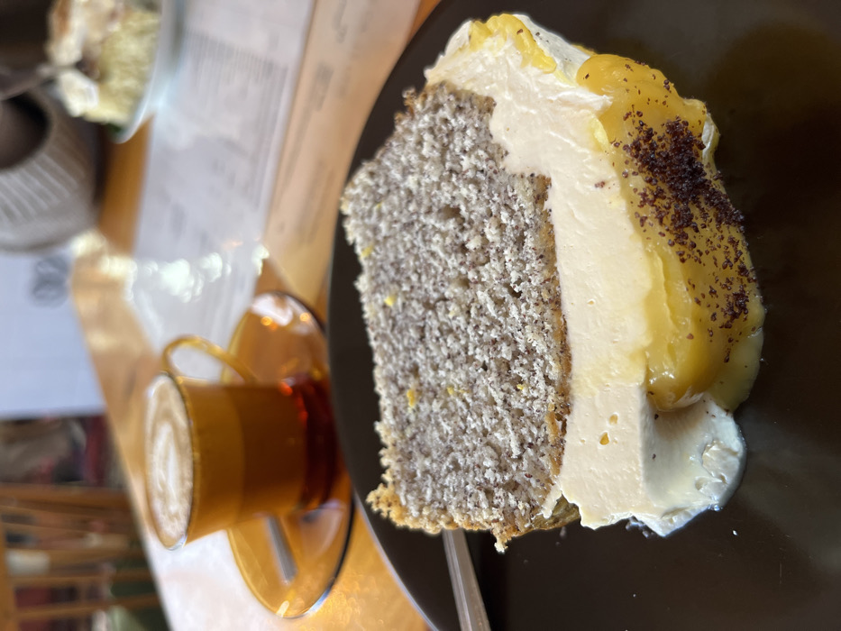 The scene depicts a dessert and beverage presented on a wooden table within an indoor cafe setting in Kaiserslautern, Germany. In the foreground, a multi-layered slice of cake rests on a dark brown plate. The cake comprises an upper grayish-brown layer densely speckled with dark particles, a central thick layer of off-white cream or frosting, and a lower vibrant yellow, semi-translucent layer consistent with fruit curd. This yellow layer partially sags onto the plate, and its bottom edge is dusted with dark brown particulate matter. A small amount of white cream has also dripped onto the plate near the cake's base. A metallic utensil handle is partially visible to the left of the cake. In the blurred midground, an amber-colored glass cup with a handle sits on a matching saucer, containing a beverage topped with white foam arranged in a spiral pattern, indicative of latte art. A small spoon rests on the saucer. The heavily blurred background reveals a light-toned wooden table surface, a folded white paper document (likely a menu), a partial view of another dessert on a plate, and indistinct elements of cafe furniture, including portions of a wooden chair and a woven decorative item. No individuals are visible.