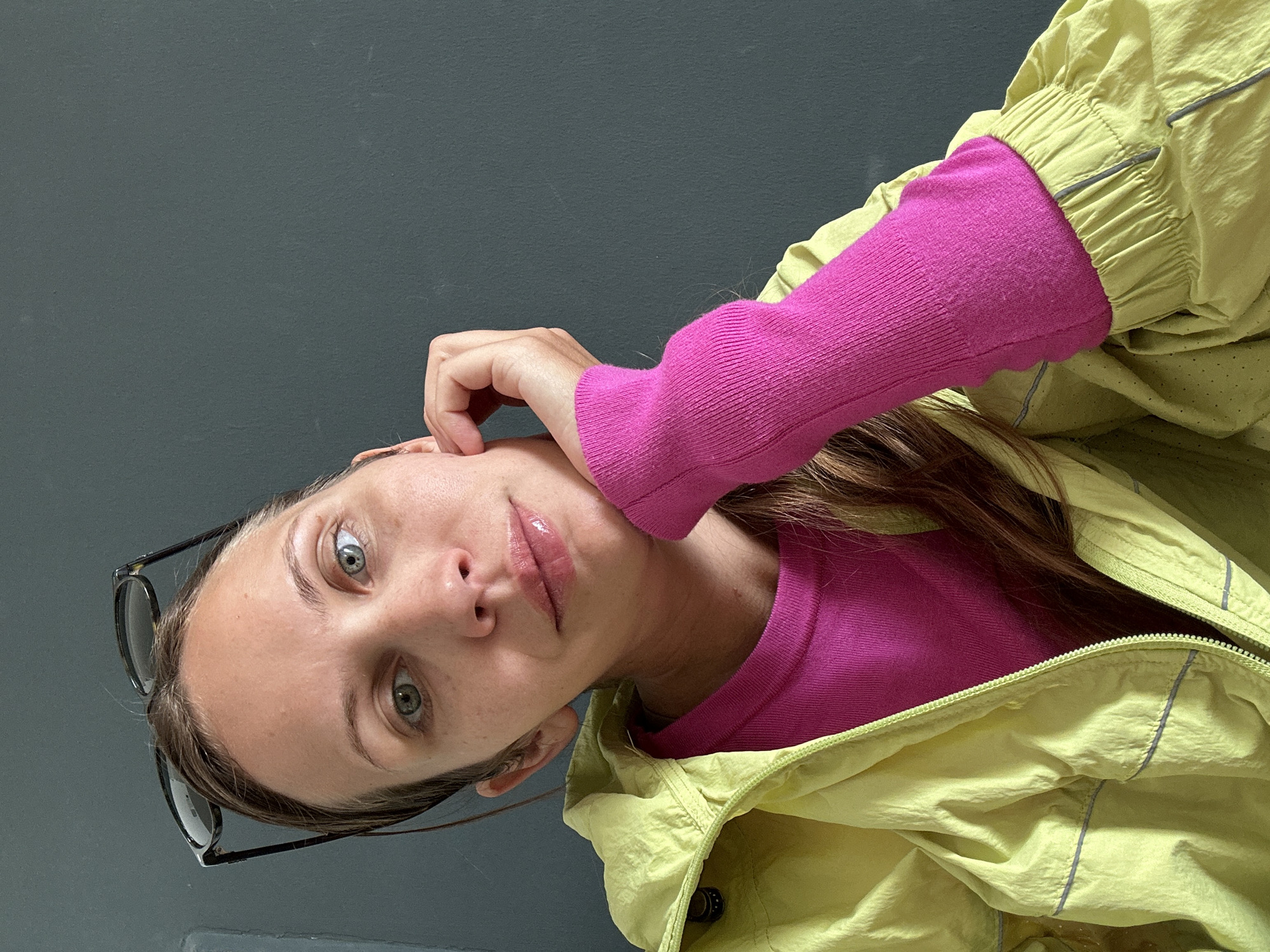 An informal, rotated, close-up photograph captures a woman, likely a selfie, looking directly at the camera. Her head is tilted, and she gazes with wide, light-colored (green/hazel) eyes, her lips slightly parted. Her right hand is raised, with fingers gently touching her ear/chin area. She has fair skin, and dark hair pulled back, with black-framed glasses resting on top of her head.
She is dressed in a vibrant, ribbed, long-sleeved fuchsia-pink top, worn underneath a lightweight, neon yellow or lime green jacket. The jacket features subtle textured details, including a zipped front and ruched elasticated cuffs. The background is a plain, dark gray wall, offering no specific location indicators within the image itself. The photograph was taken in Fethiye, Turkey. The lighting appears soft and even, suggesting an indoor setting or shaded outdoor area. There is no visible text in the image.