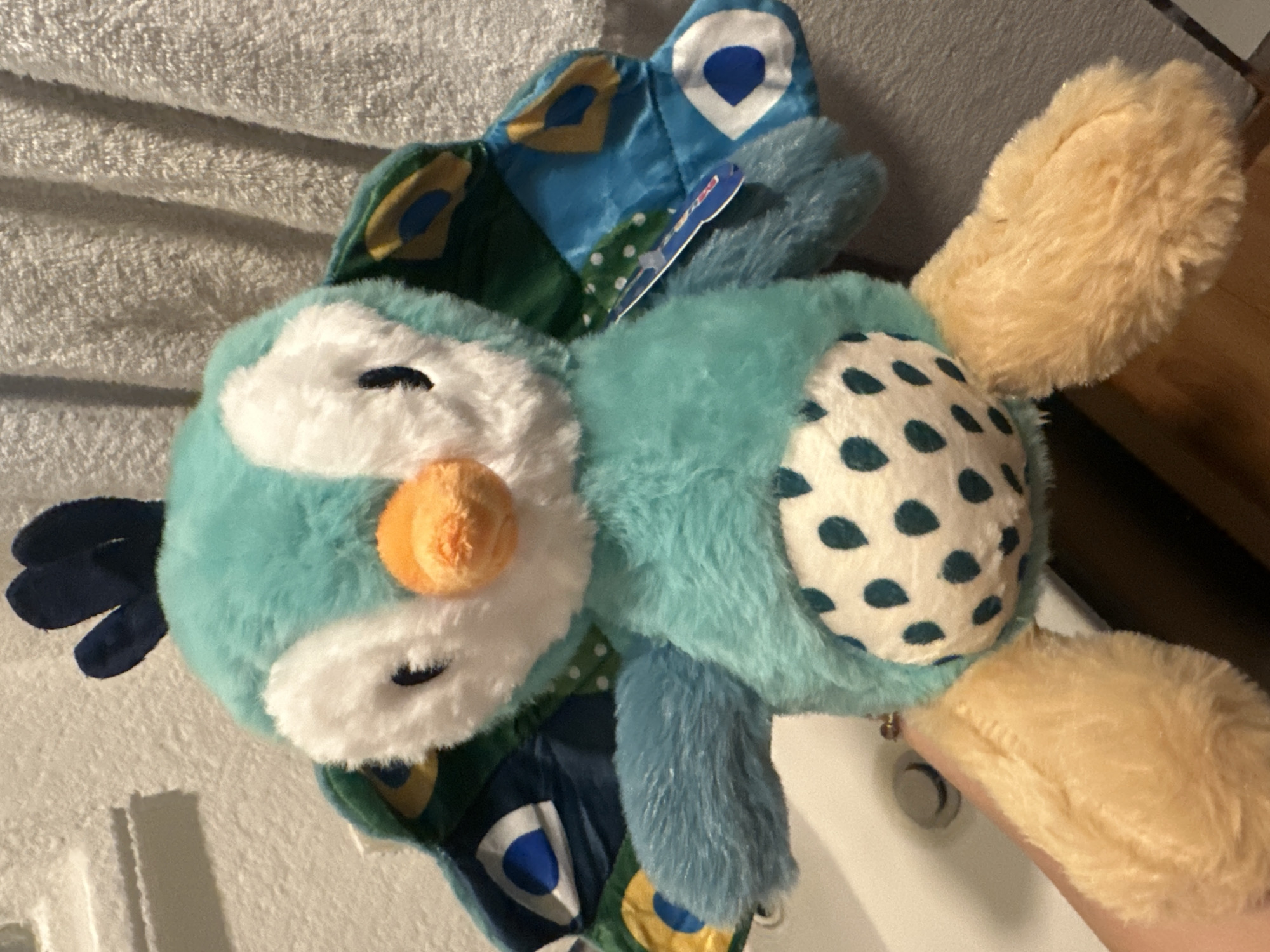 A colorful owl plush toy is positioned atop a white, textured surface. The toy features a blue and white face with a prominent orange nose. It has a patterned back in white with dark teal spots and tan-colored wings. The owl also has a section of peacock-like feathers with blue, green, and gold colors. A tag is visible near the center of the toy. The overall composition is soft and inviting, suggesting a child's toy or decoration. The location is not discernable from this image.