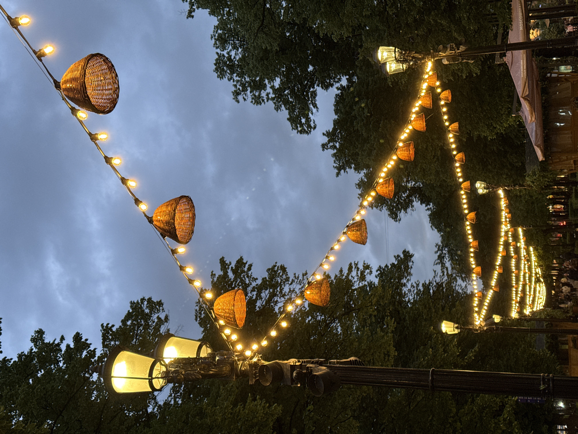 The image captures a street scene in Moscow, Russia. A string of decorative lights hangs diagonally across the frame, featuring alternating wicker baskets and small light bulbs. The lights are suspended above a street lined with trees. The sky is overcast. Traditional street lamps are visible at the bottom left and along the right side of the frame.