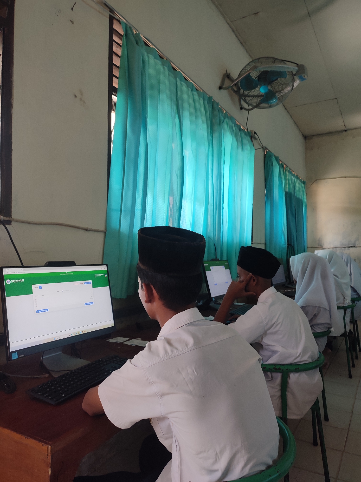 In what appears to be a computer laboratory in Blangriek, Indonesia, multiple students are seated at computer workstations. The students wear white shirts and black peci hats, and one student is wearing a white hijab. The students are focused on the computer screens in front of them. A computer monitor displays the logo of "Garuda CBT" and a data entry form. The setting is a simple classroom with desks and chairs. A light blue curtain hangs on the window in the background. A wall-mounted fan is visible. The tiled floor suggests a school or similar educational setting.