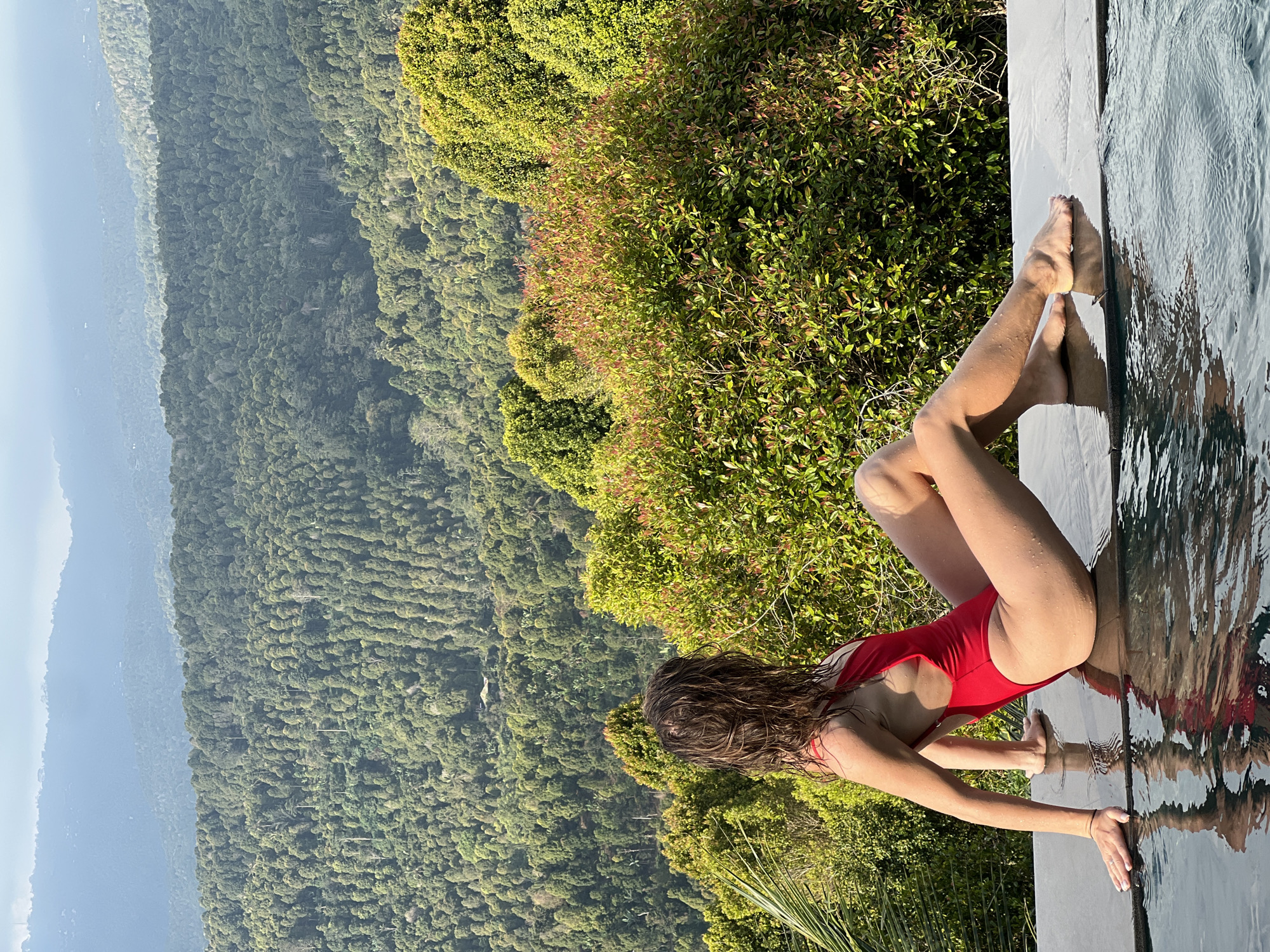 In Busungbiu, Indonesia, a woman in a red one-piece swimsuit sits on the edge of an infinity pool, her back to the camera. Her wet, brown hair falls over her shoulders. The pool's edge is made of dark, flat stones, and the water's surface reflects the scene. Beyond the pool, a lush, green landscape unfolds, with dense forests covering rolling hills and mountains stretching into the distance. Brightly lit green and reddish bushes are visible in the middle ground, bordering the pool. The sky is hazy and light blue.