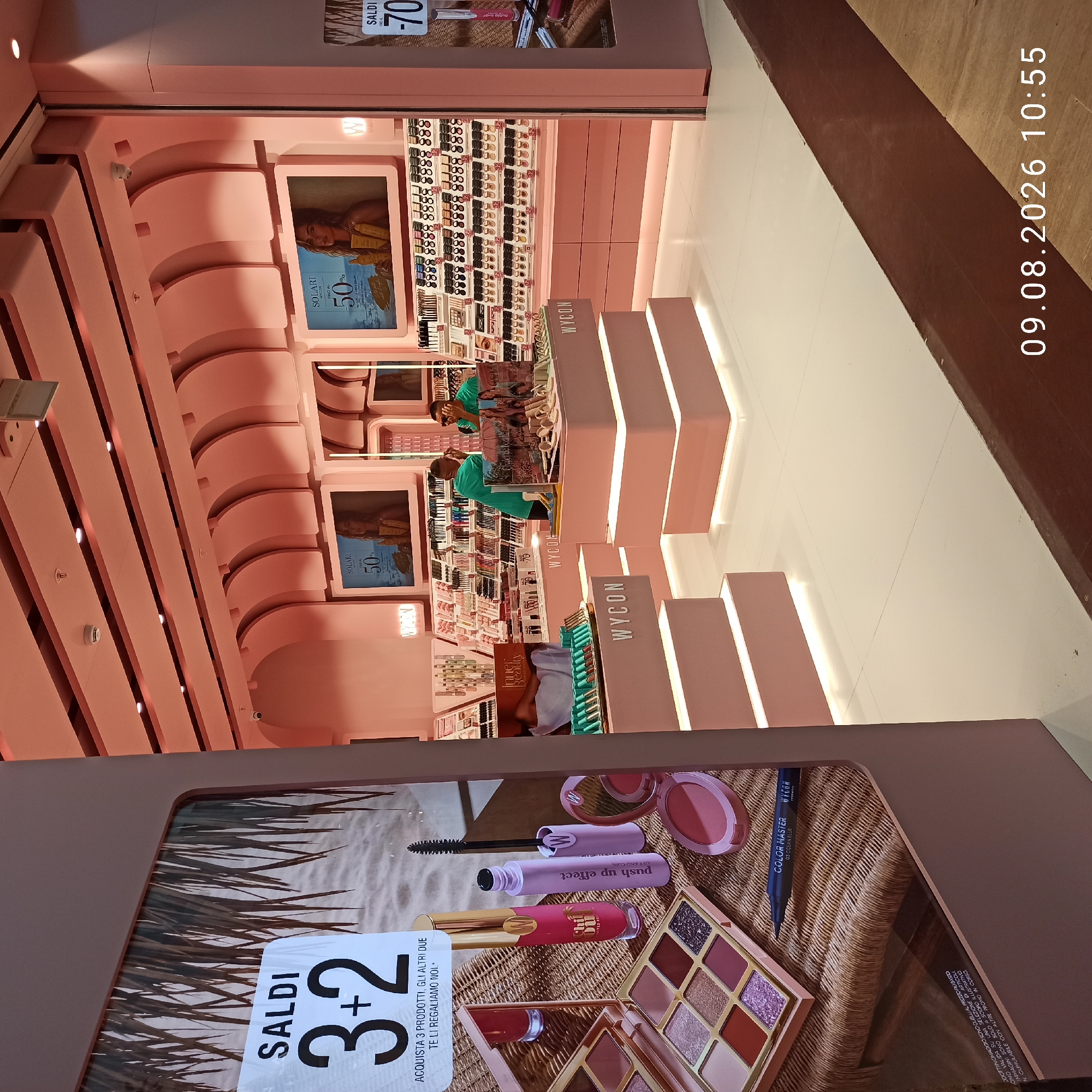 The image depicts the interior of a Wycon cosmetics store, a brand commonly found in Italy. The store's aesthetic is characterized by predominantly pink decor, with illuminated shelving units displaying various makeup products. In the foreground, a large promotional poster is visible, advertising a "SALDI 3+2" offer, which translates to "SALE 3+2," indicating a "buy 3, get 2 free" promotion. The poster features images of makeup items, including eyeshadow palettes, lipstick, and mascara, set against a beach-like background.
Further into the store, several display stands are arranged. One prominent display features rows of makeup products, possibly eyeshadows and blushes. Another display showcases an array of lipstick or lip gloss tubes and mascara. Two individuals are present in the background, one wearing a green t-shirt and the other in a similar colored shirt, appearing to be store employees interacting with the merchandise or assisting customers. One of the individuals is wearing sunglasses.
Several screens are mounted on the pink walls, displaying advertisements. One screen shows a model and text related to "SOLARI" (sunscreens) with a "50%" discount. Another screen displays "SALDI -70%". The overall environment suggests a retail setting focused on beauty products, with clean lines and bright lighting, enhancing the display of the merchandise. The timestamp "09.08.2026 10:55" is superimposed on the lower right corner of the image, indicating the date and time the photograph was taken.
While the image itself does not explicitly state the location within Molfetta, Italy, the presence of a Wycon store, an Italian brand, strongly suggests it is within the city or its immediate vicinity.