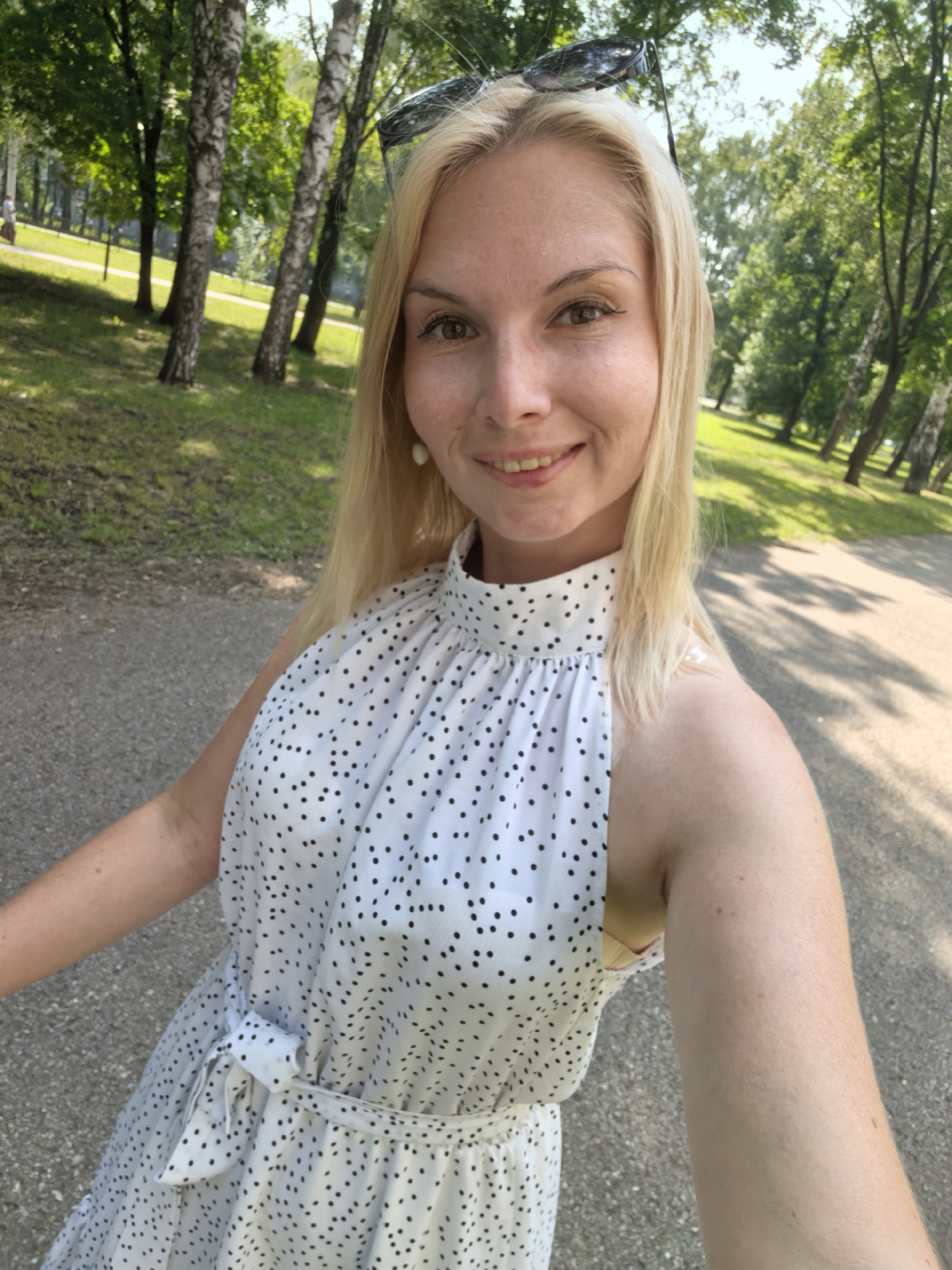 A blonde woman is taking a selfie in a park. She is wearing a white polka dot dress with a high neckline. Sunglasses are perched on top of her head. She is smiling at the camera. The background shows trees and a paved path. It is a sunny day.