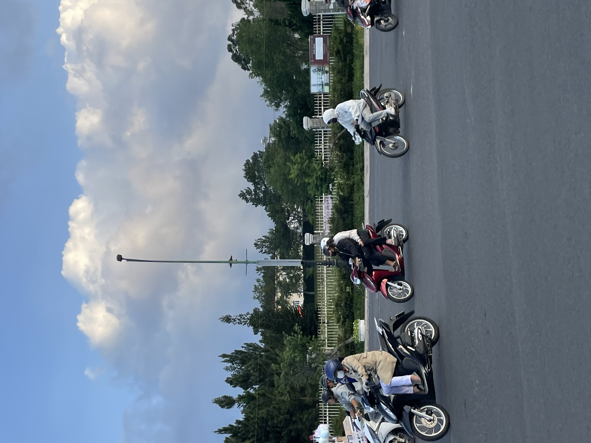 The image depicts a street scene in Da Lat, Vietnam, under a sky with prominent white cumulus clouds against a light blue backdrop, suggesting late afternoon or early evening. The foreground is a wide, dark gray asphalt road.
Six individuals are visible, all operating or riding on motorbikes or scooters. From left to right:
1. A white scooter, partially visible on the far left, has a rider in a dark helmet and clothing, with an object strapped to the front.
2. A person wearing a dark helmet, a light-colored face mask, and a long tan coat over white pants is riding a black Honda Vision scooter with silver-spoked wheels.
3. A red Honda Vision scooter is ridden by two individuals. The driver wears a dark helmet and dark clothing. The passenger wears a white hooded top and dark pants, with a face mask.
4. A person wearing a white hooded jacket, a white face mask, light gray pants, and white sneakers is riding a black underbone-style motorbike with silver-spoked wheels.
5. A dark-colored motorbike is partially visible on the far right, with a rider in dark clothing.
In the mid-ground, a tall, slender dark green or black lamppost stands. Behind the lamppost and motorbikes, a white ornamental metal fence separates the road from a densely vegetated area featuring numerous tall, slender pine trees and other foliage. Beyond the trees, white buildings are partially visible, and a large sign with Vietnamese text is present on the right side of the fence.