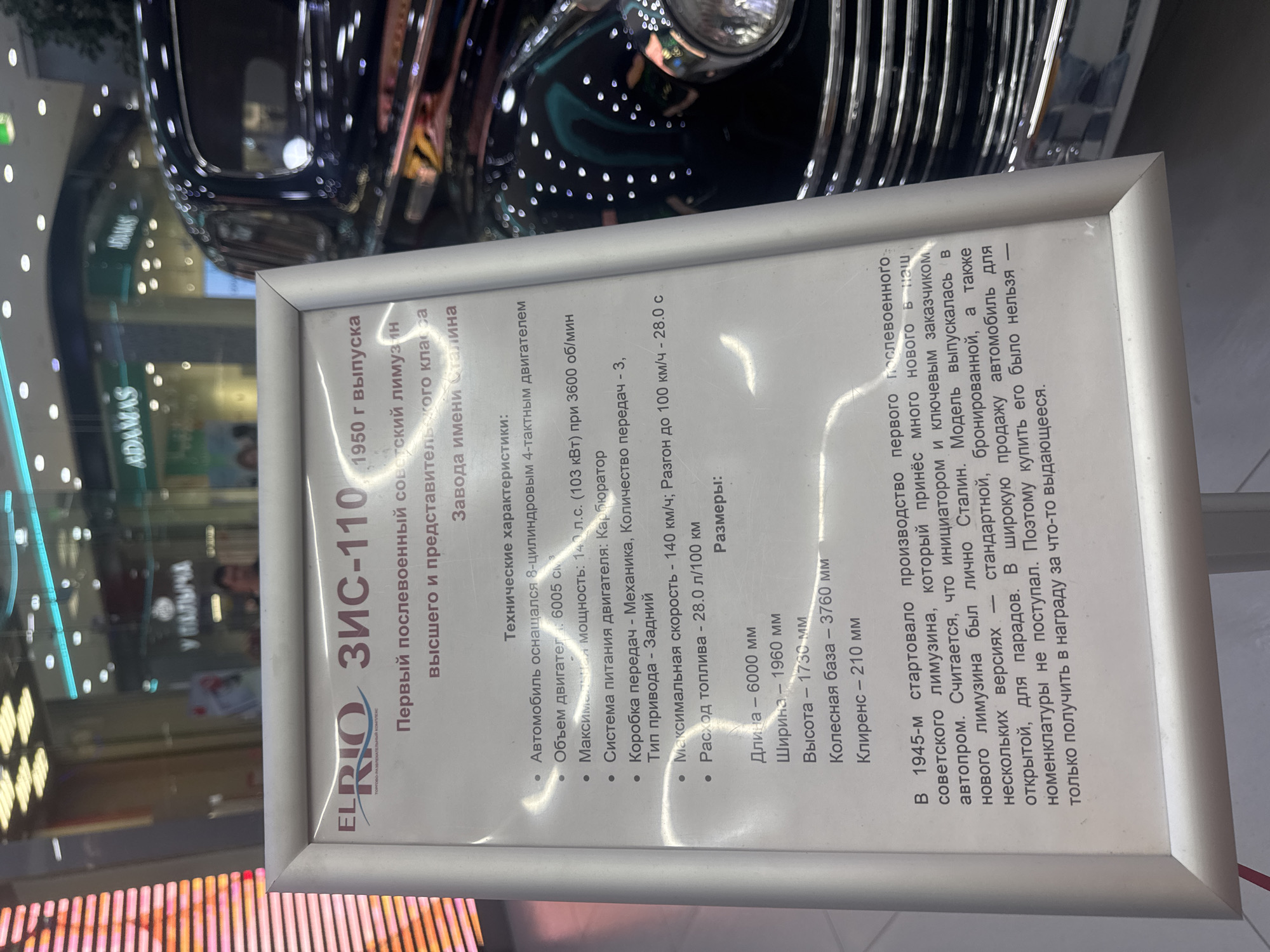 This media file captures a historical display, likely within a museum or exhibition space. The central focus is a plaque detailing the ZIS-110, a Soviet limousine from 1950. The plaque, framed in silver, is positioned in front of a black vintage car, only partially visible, hinting at its impressive size and luxurious design. The text on the plaque is in Russian and provides technical specifications of the car, including engine size, power, maximum speed, and dimensions. It also offers historical context, mentioning that the ZIS-110 was a symbol of prestige and was even associated with Joseph Stalin. The background shows a blurred retail or exhibition environment, with reflections of lights and store signage, suggesting an indoor setting within a public space. The overall atmosphere is informative and nostalgic, highlighting a significant piece of Soviet automotive history.